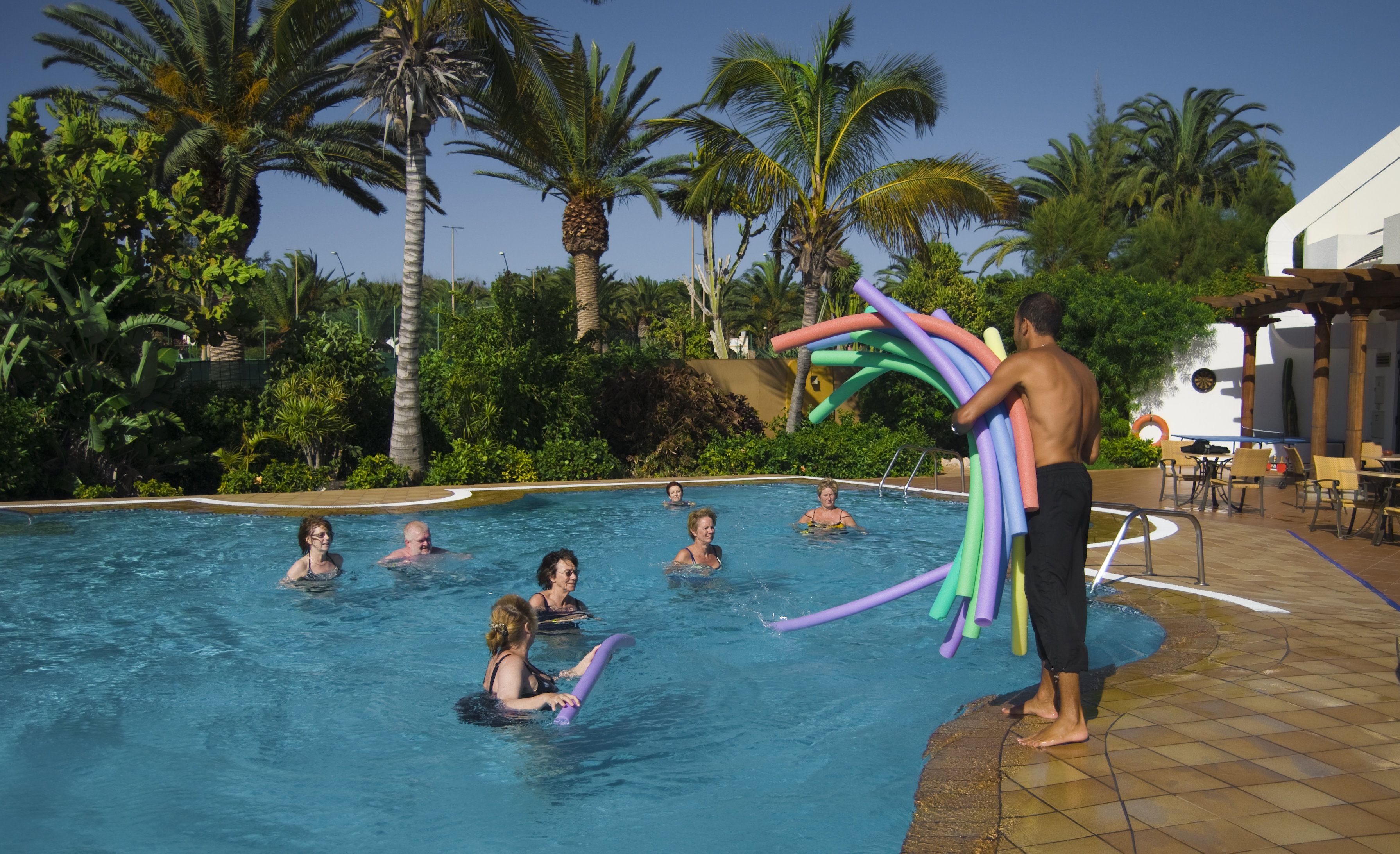 Une piscine où des personnes se baignent, entourée de palmiers et d’un espace patio aménagé avec des chaises et des tables.