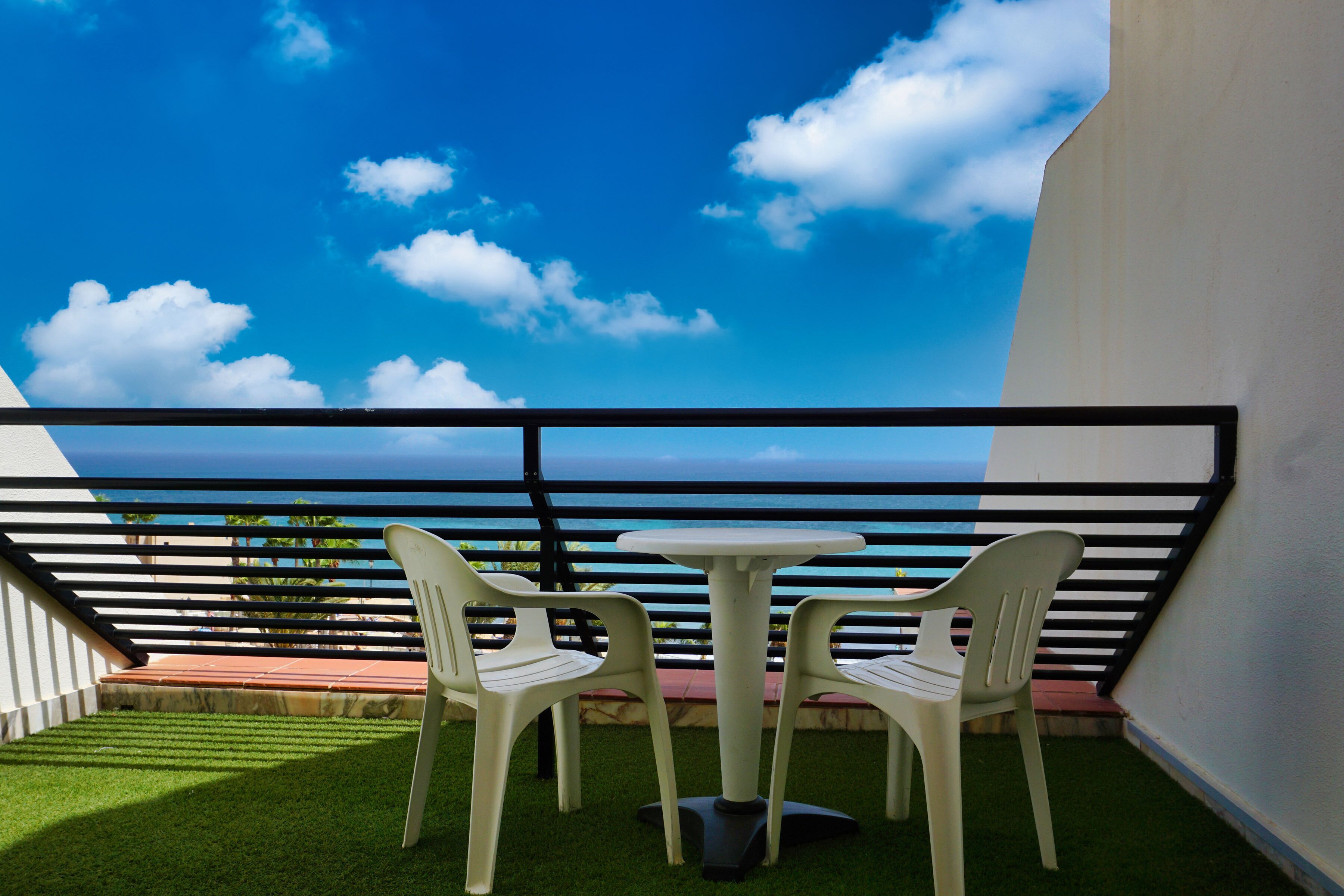 Ein Balkon mit zwei weißen Plastikstühlen und einem kleinen runden Tisch, mit Blick auf eine schöne Aussicht auf das Meer und Wolken.