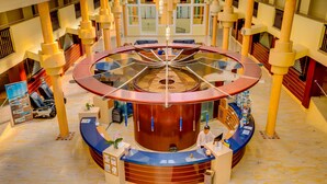 Un hall de hotel con un mostrador de recepción circular central, varias áreas de descanso y un techo alto con luces colgantes.