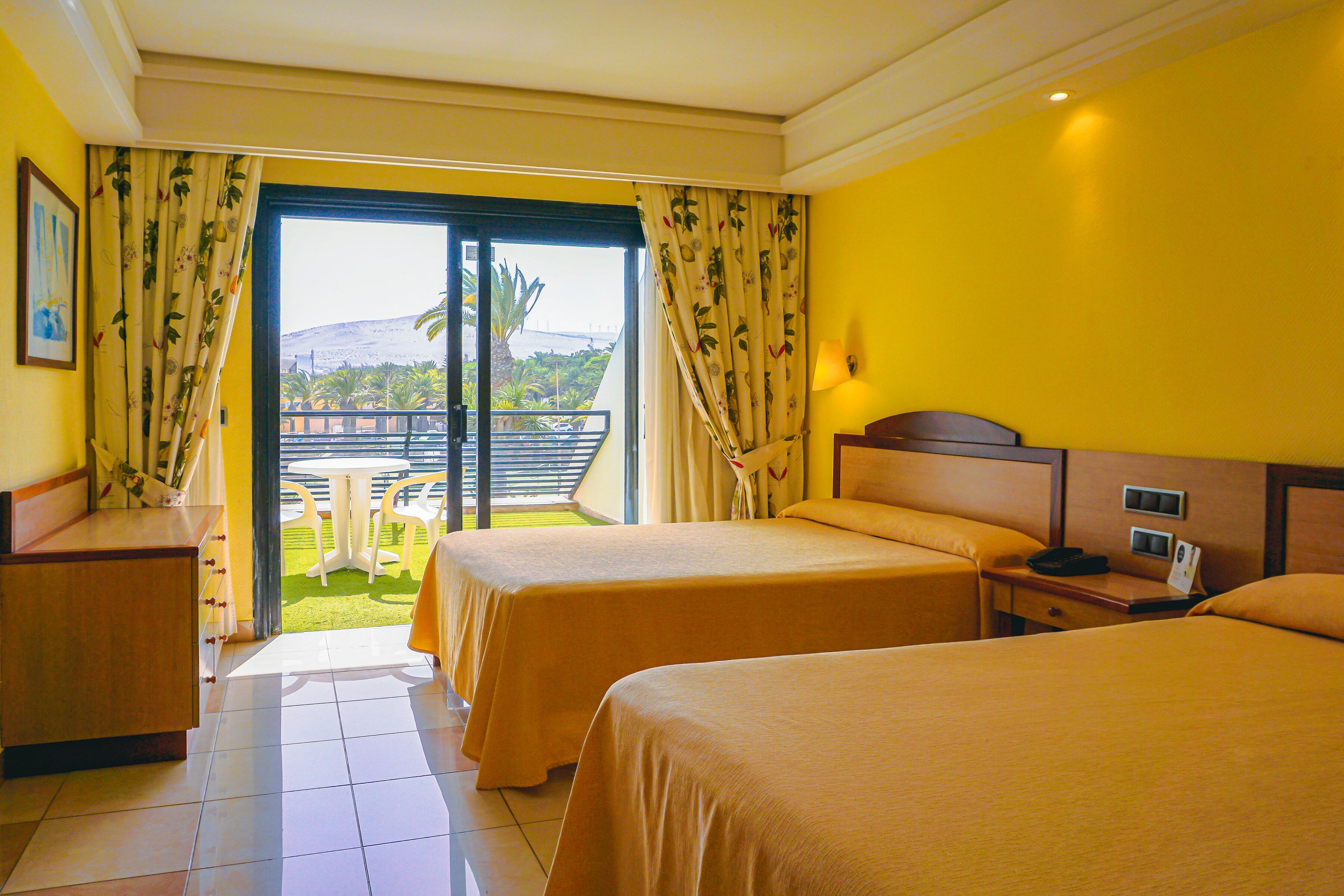 A hotel room with two beds, a desk, a chair, and a balcony with a view of the landscape.