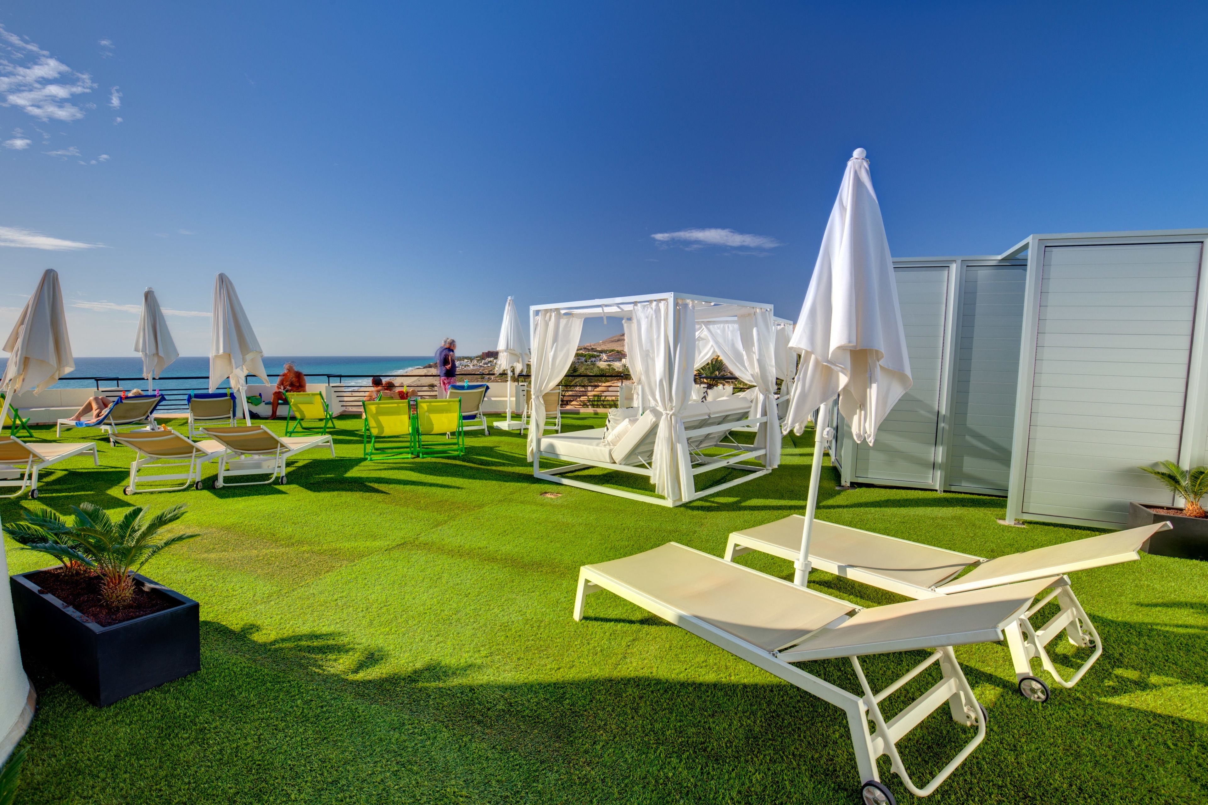 Un espace de détente aménagé au bord de la plage, avec des chaises longues et des parasols blancs, une vue dégagée sur la mer et une pelouse verdoyante bien entretenue.