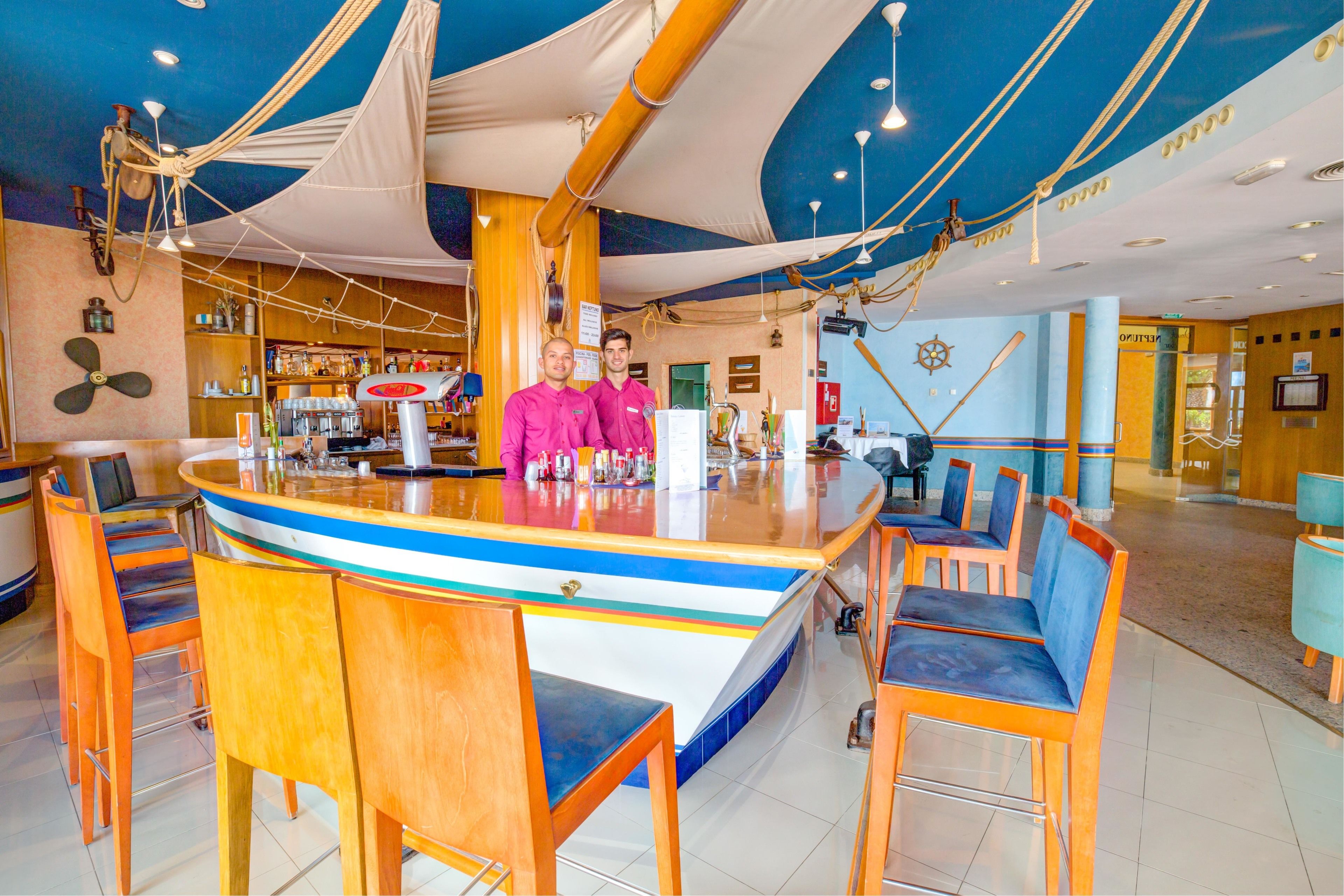 Un espace bar avec des chaises en bois et un comptoir, agrémenté d’une décoration sur le thème de la marine, comprenant des cordes et un gouvernail de navire.
