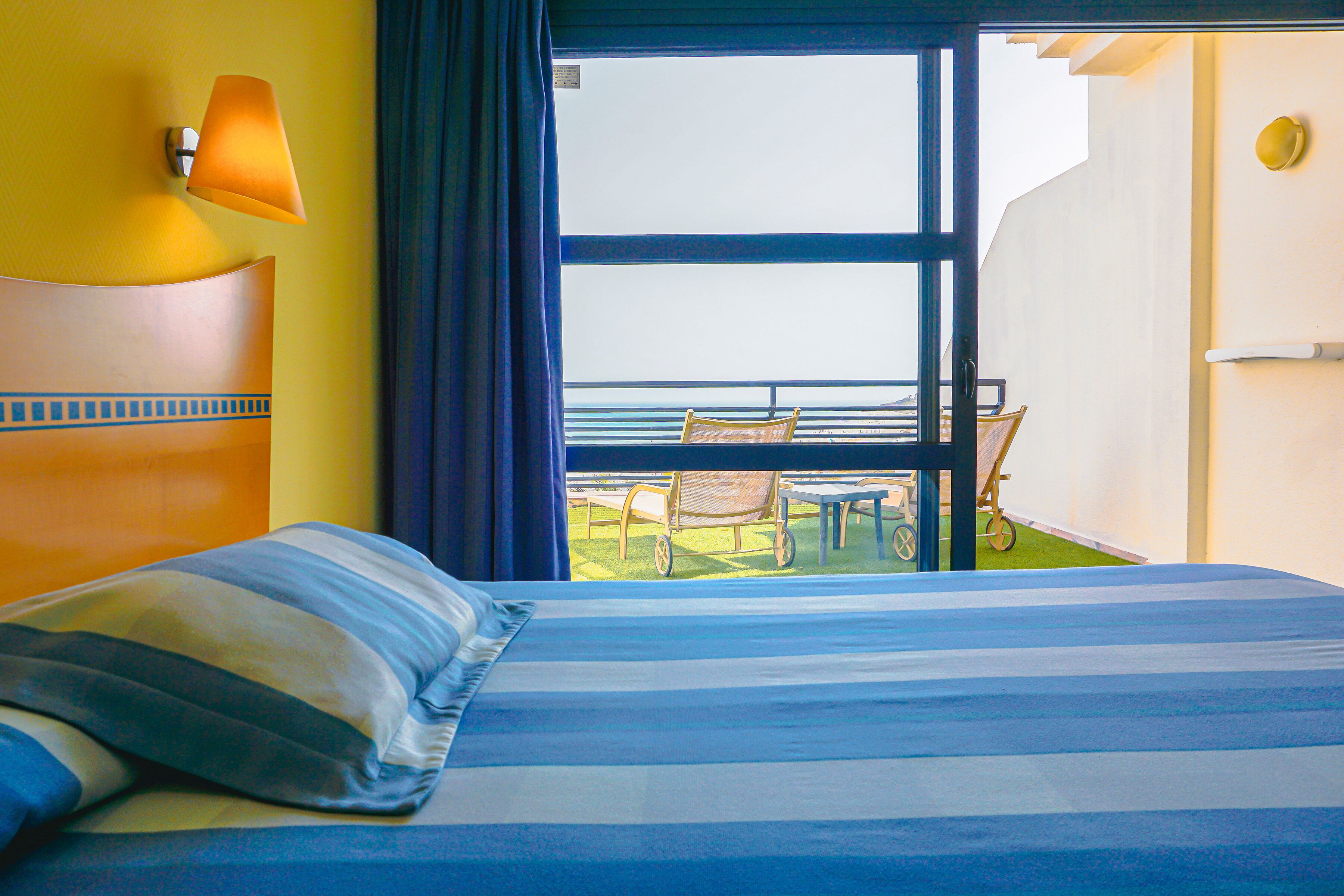 A hotel room with a large bed, a view of the sea, a balcony with outdoor furniture, and a lamp on the wall.