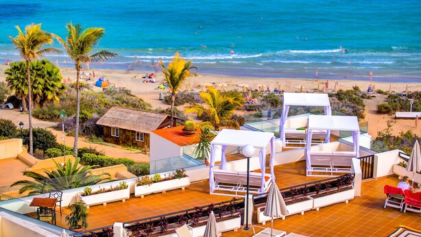 Terraza en la azotea con vistas a la playa, palmeras y un edificio pequeño.