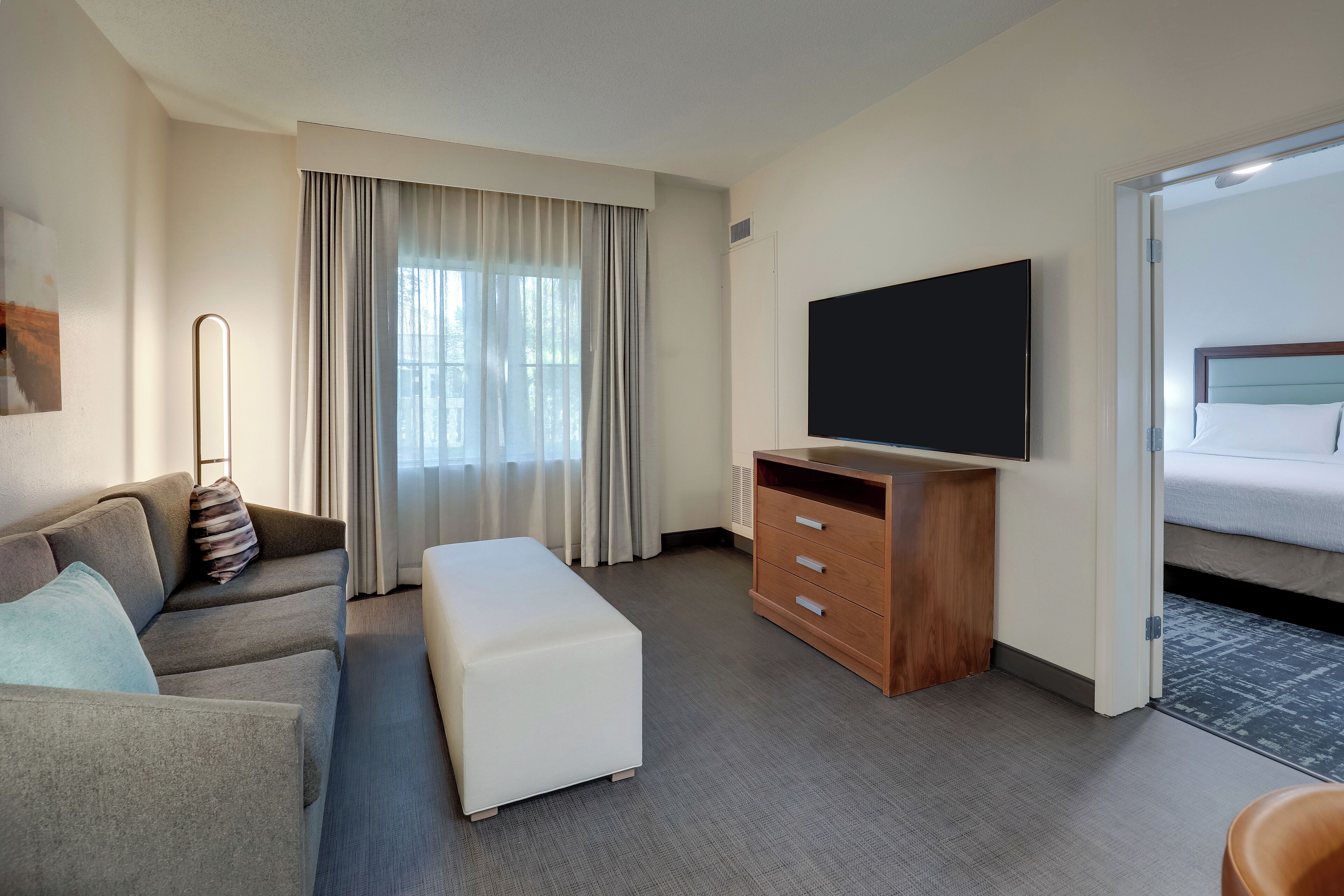 A hotel room with a sofa, ottoman, wooden dresser, flat-screen TV, and a bed.