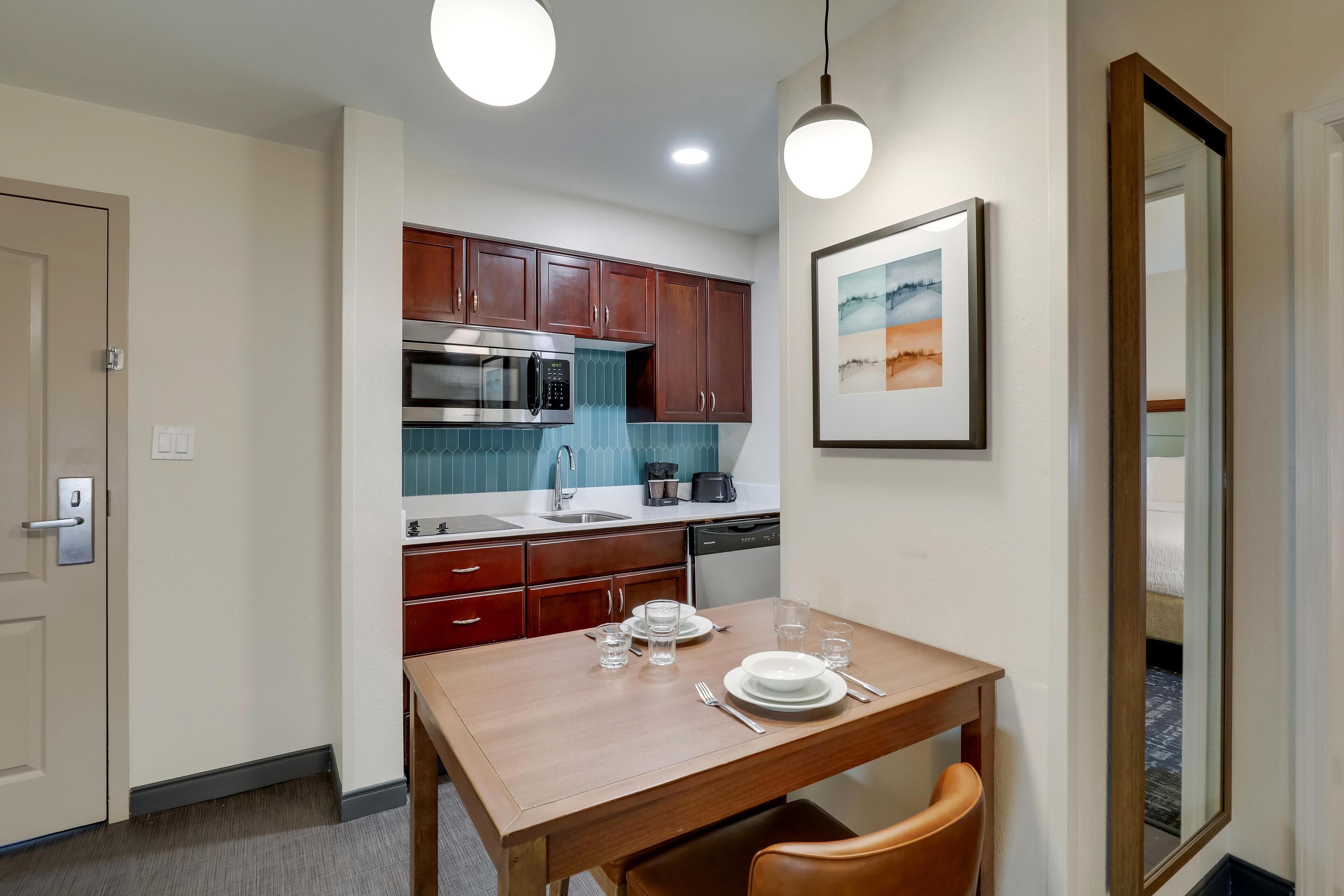 A compact kitchen with wooden cabinets, a stainless steel microwave, and a dishwasher. There is a dining table set with plates, glasses, and cutlery.