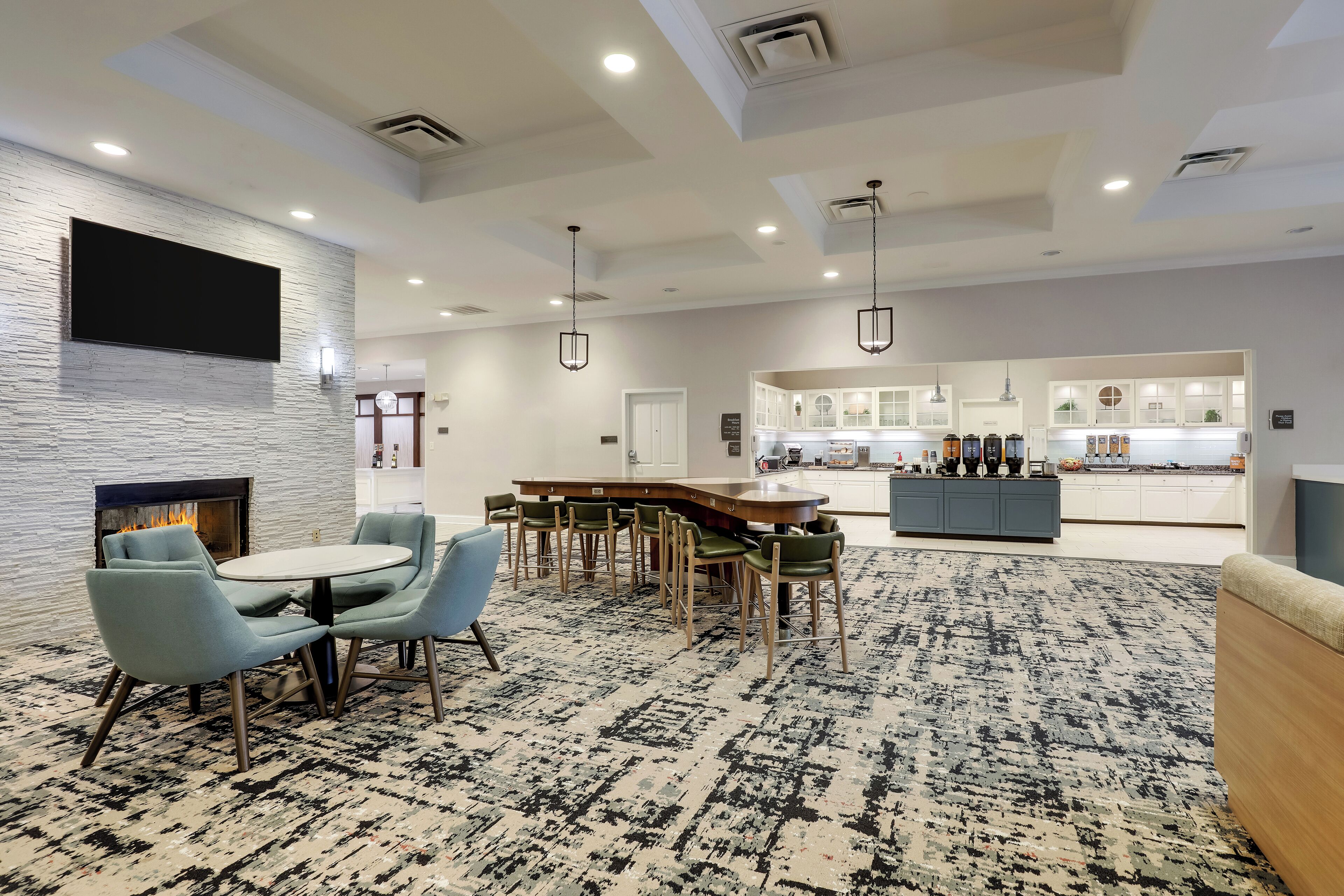 A modern hotel lobby with a fireplace, a round dining table with chairs, a kitchen area, and a patterned carpet.
