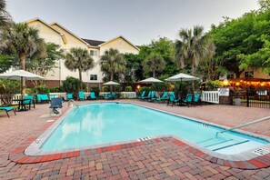 A swimming pool with lounge chairs and umbrellas, surrounded by palm trees and a brick patio.