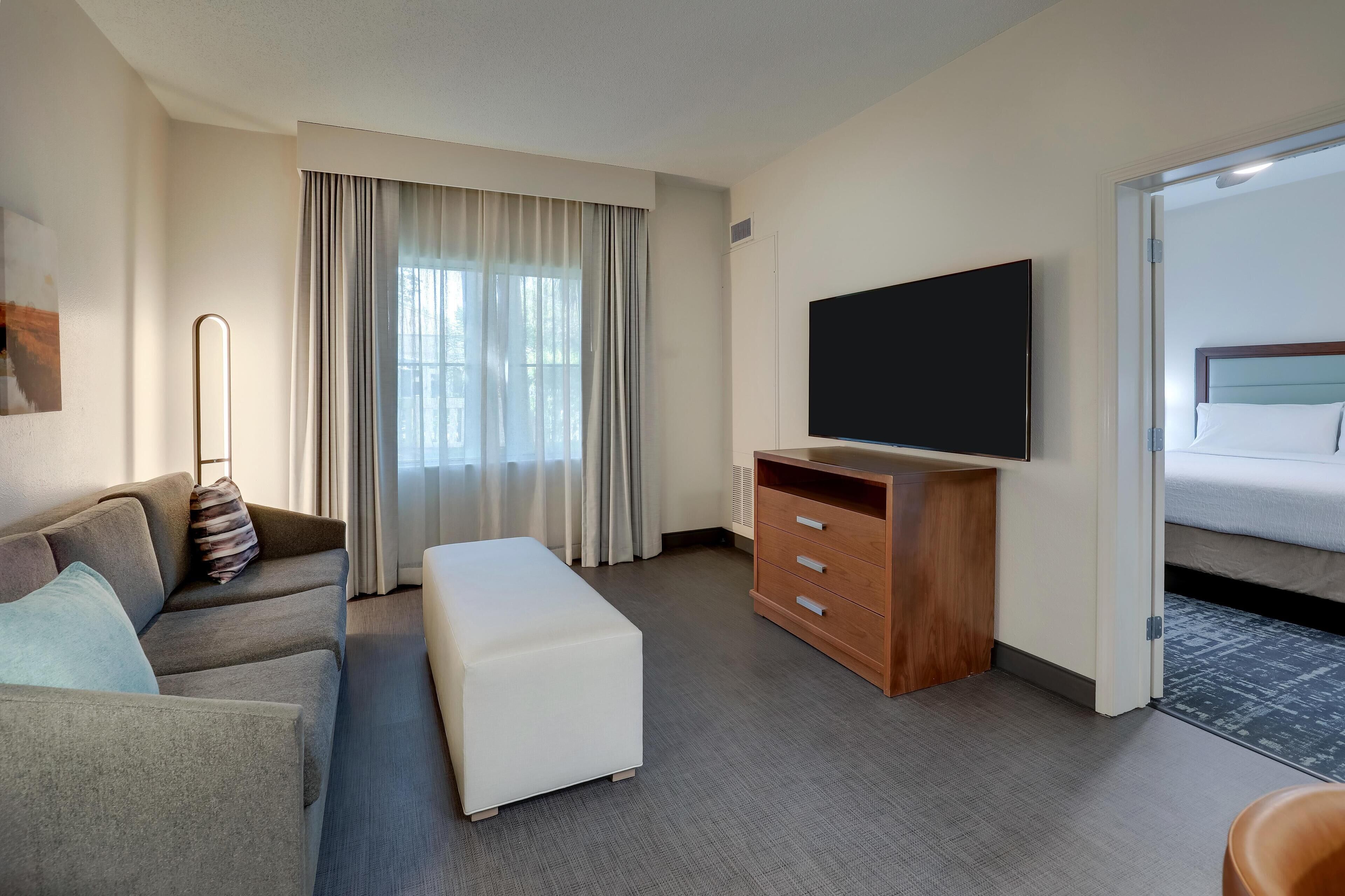 A hotel room with a sofa, ottoman, TV, wooden dresser, and a bed.