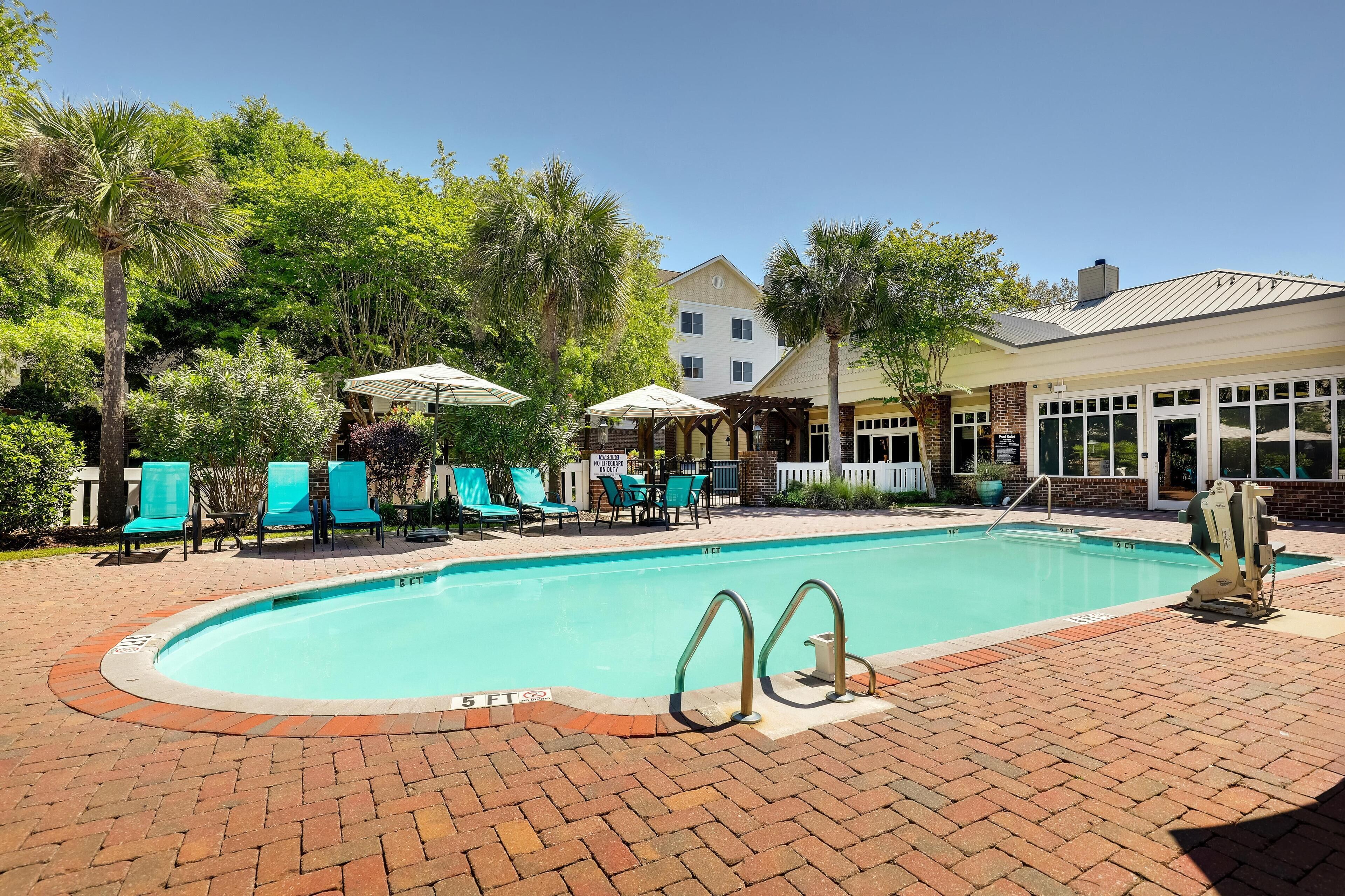 A swimming pool with a brick deck, surrounded by lounge chairs and umbrellas, and a building with large windows in the background.
