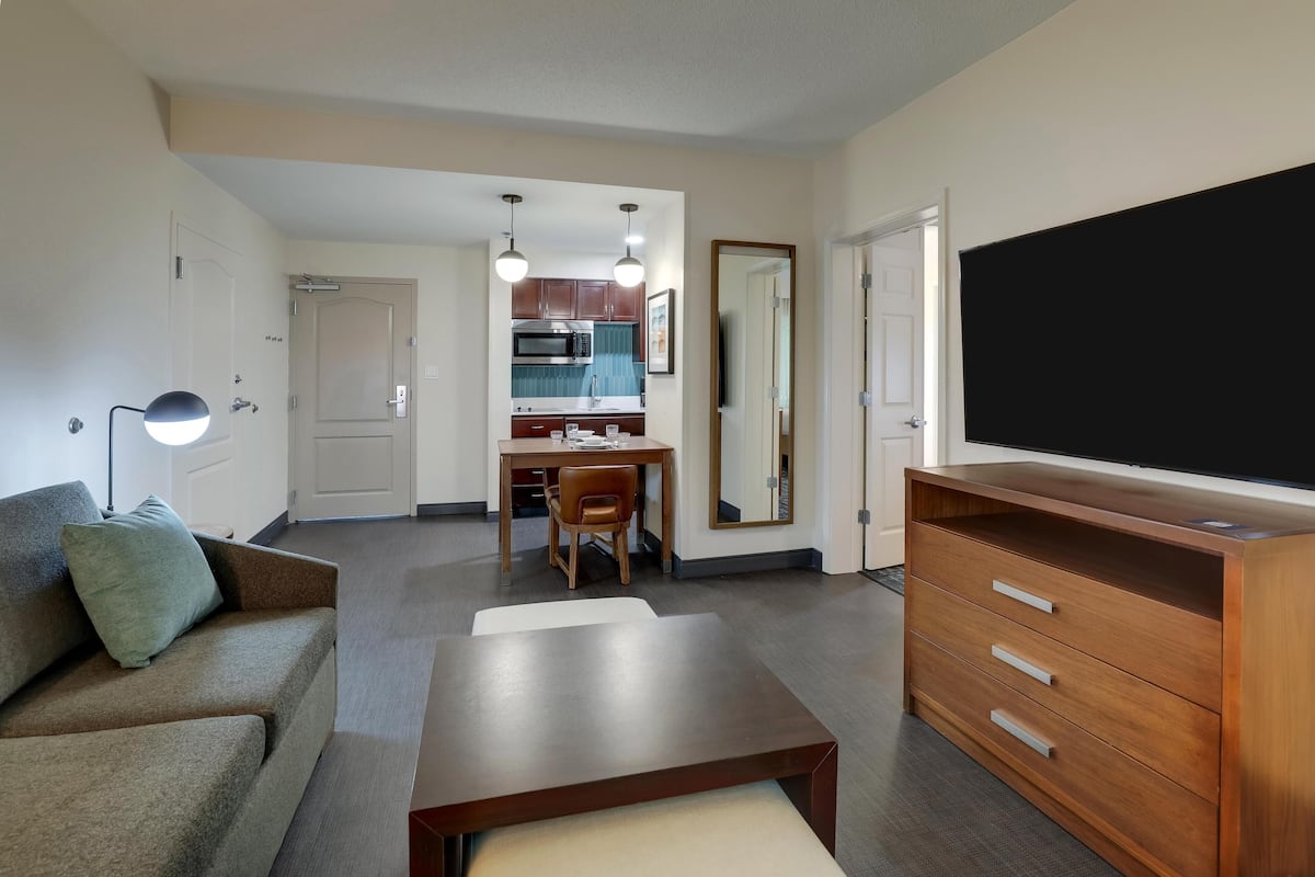 Een ruimtelijk ingerichte leefruimte met een bank, een houten dressoir, een televisie, een eettafel met stoelen en een kitchenette op de achtergrond.