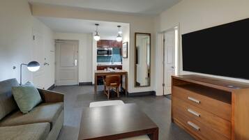Un espacio habitable compacto con sofá, cómoda de madera, televisor, mesa de comedor con sillas y una kitchenette al fondo.