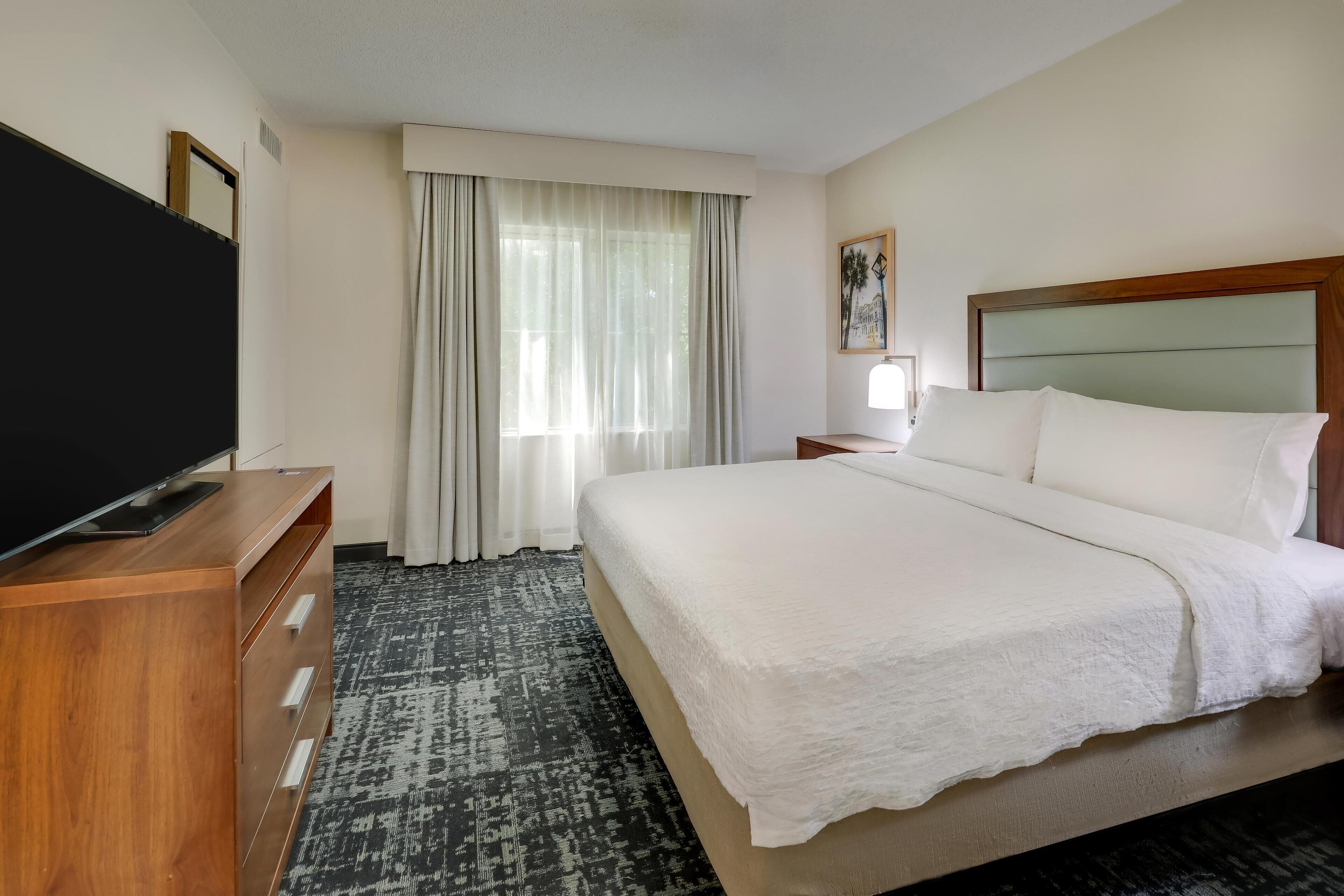 A hotel room with a large bed, a wooden dresser with a flat-screen TV, a window with sheer curtains, and a framed picture on the wall.