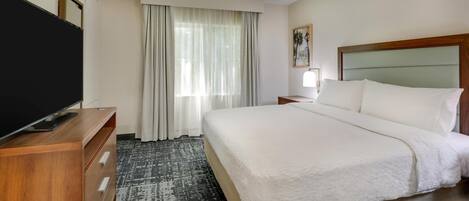 A hotel room with a large bed, a wooden dresser with a flat-screen TV, a window with sheer curtains, and a framed picture on the wall.