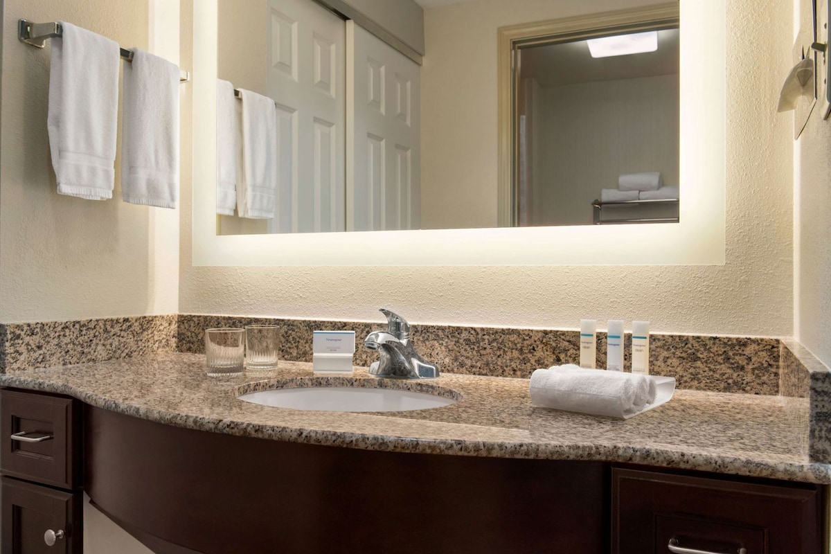 Un baño con encimera de granito, un lavabo, un espejo y un perchero con toallas blancas.