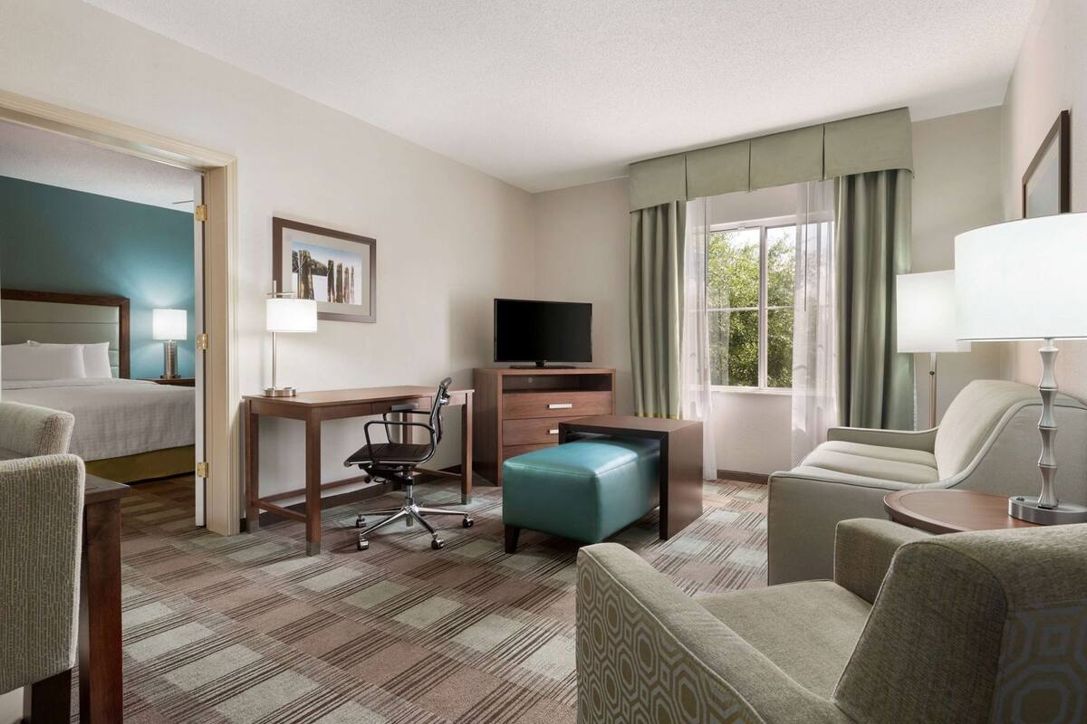 Une chambre d’hôtel comprenant un lit, un bureau, une chaise, un pouf, une télévision et une fenêtre avec des rideaux.