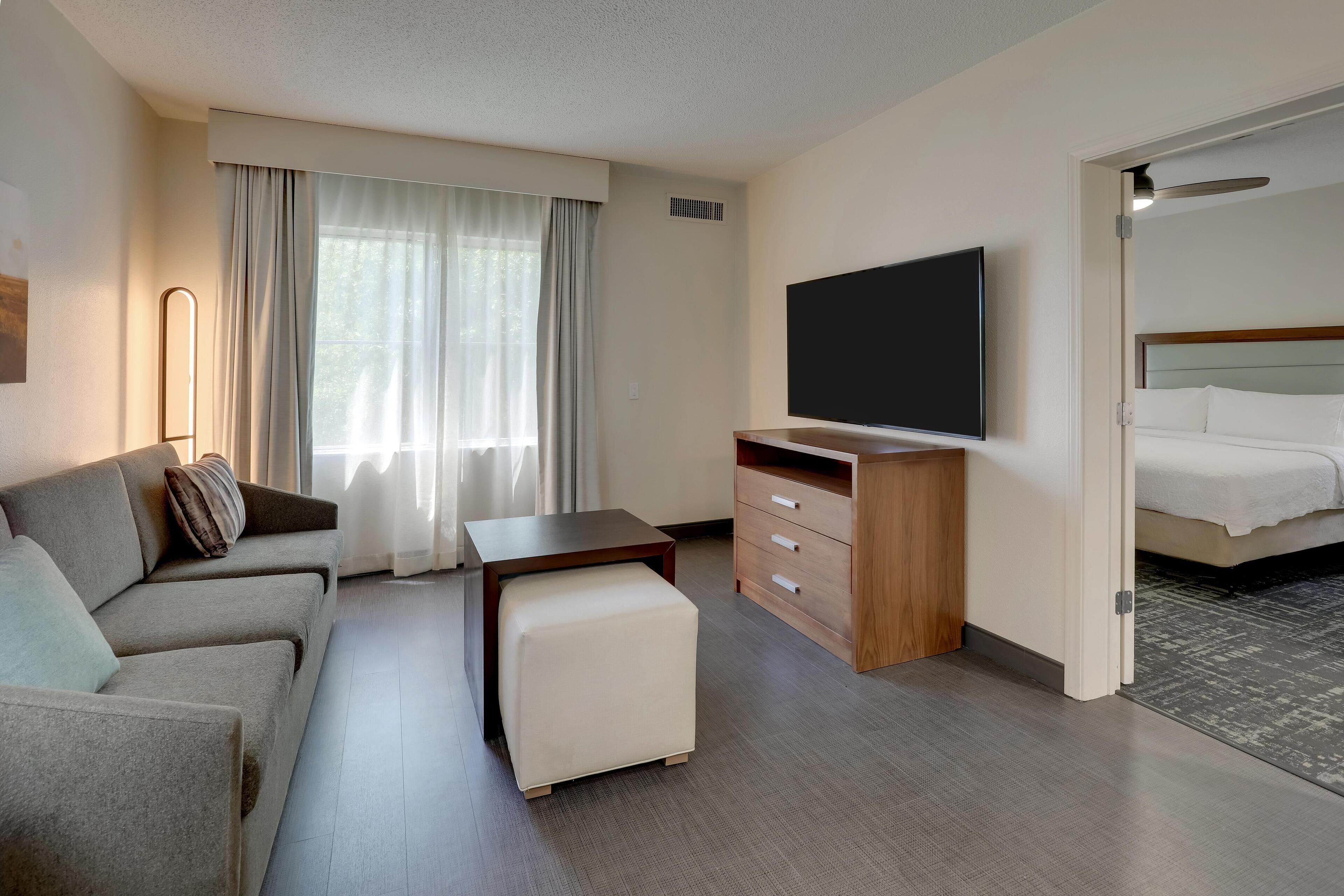 Une chambre d’hôtel moderne équipée d’un canapé, d’un pouf, d’une télévision, d’un bureau en bois et d’un lit avec une tête de lit.