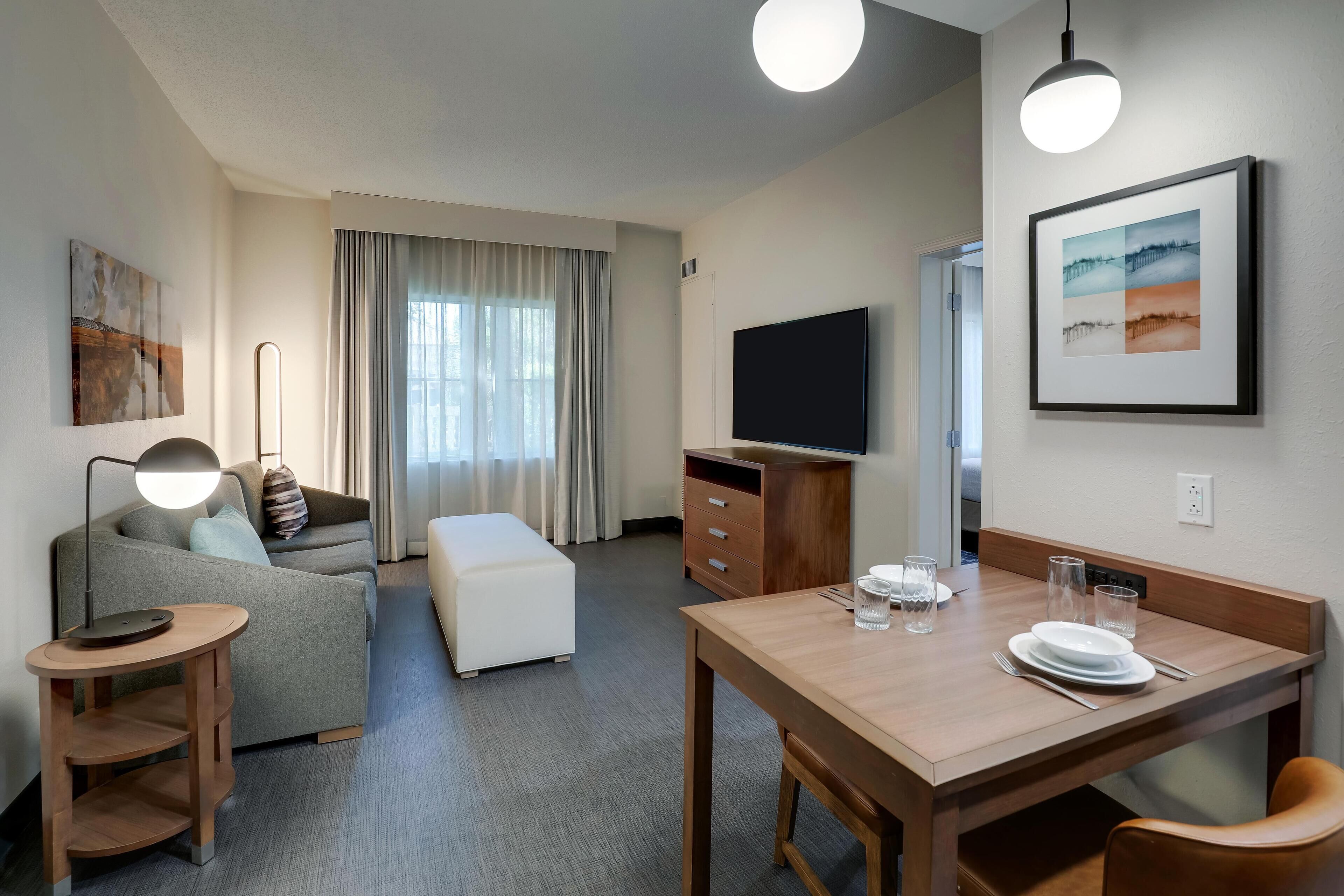 A modern hotel room with a sofa, a dining table set for two, a flat-screen TV, and a large window with curtains.