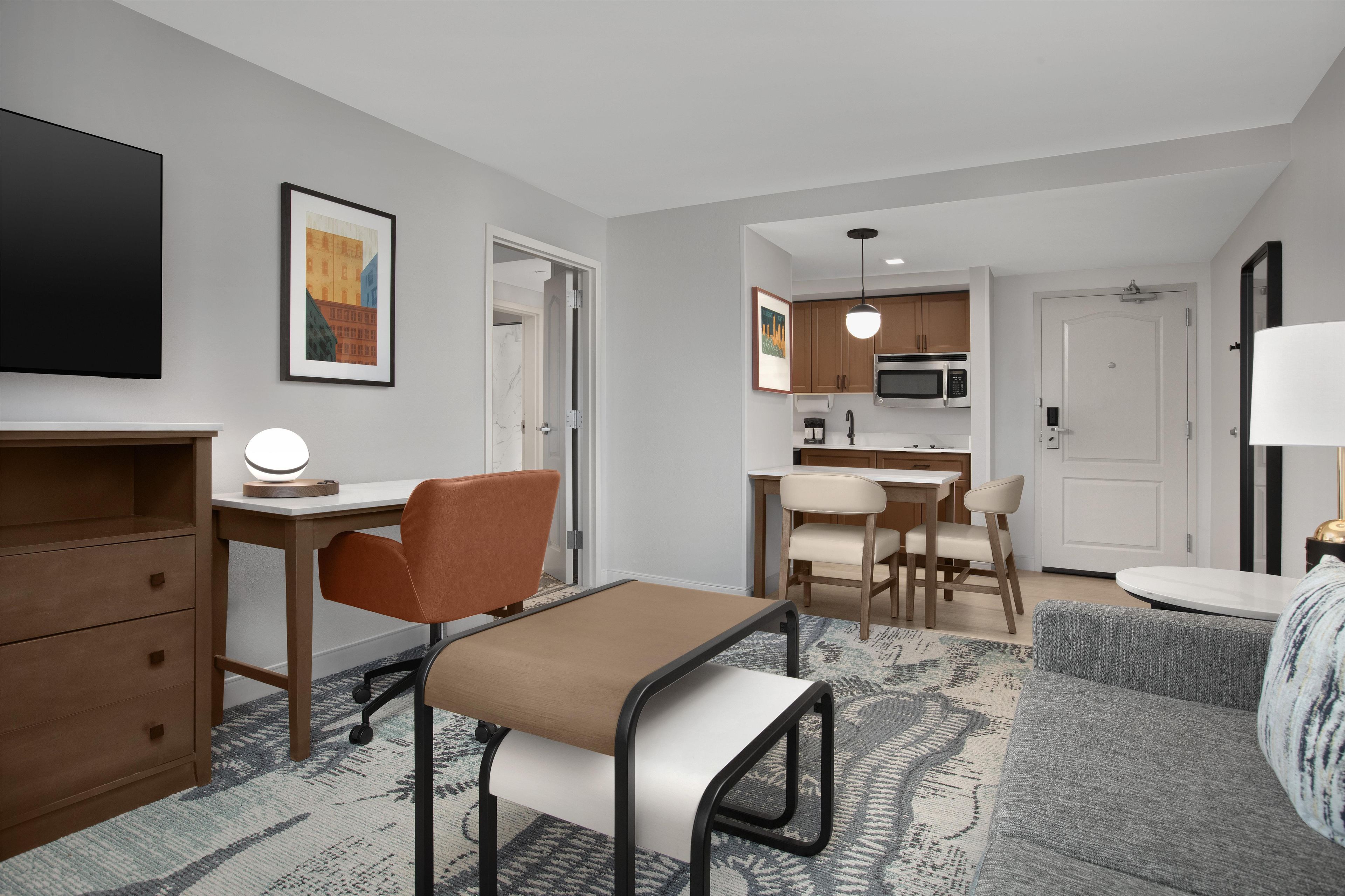 A modern hotel room with a desk, chair, sofa, and kitchenette.