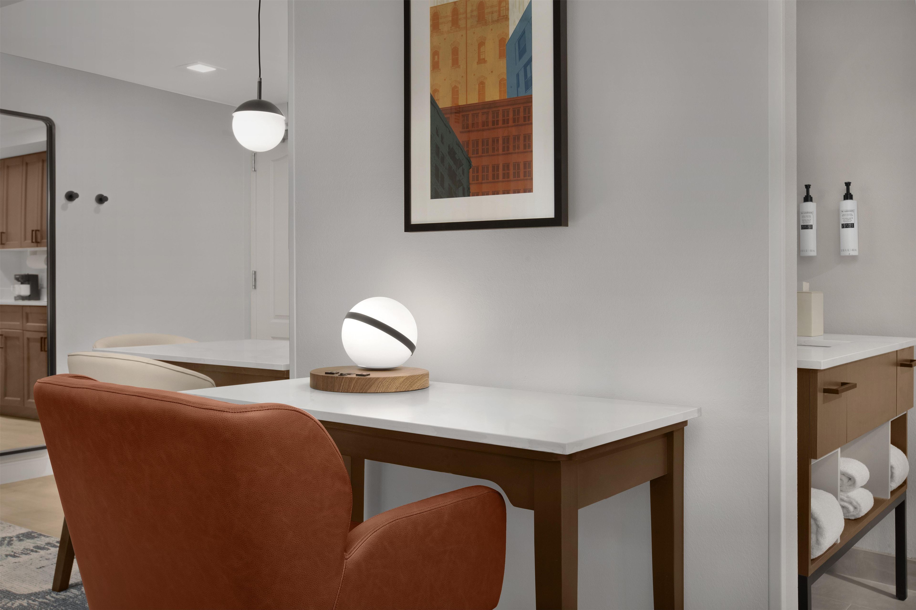 A modern room with a white desk, a brown chair, a round table lamp, and a framed picture on the wall.