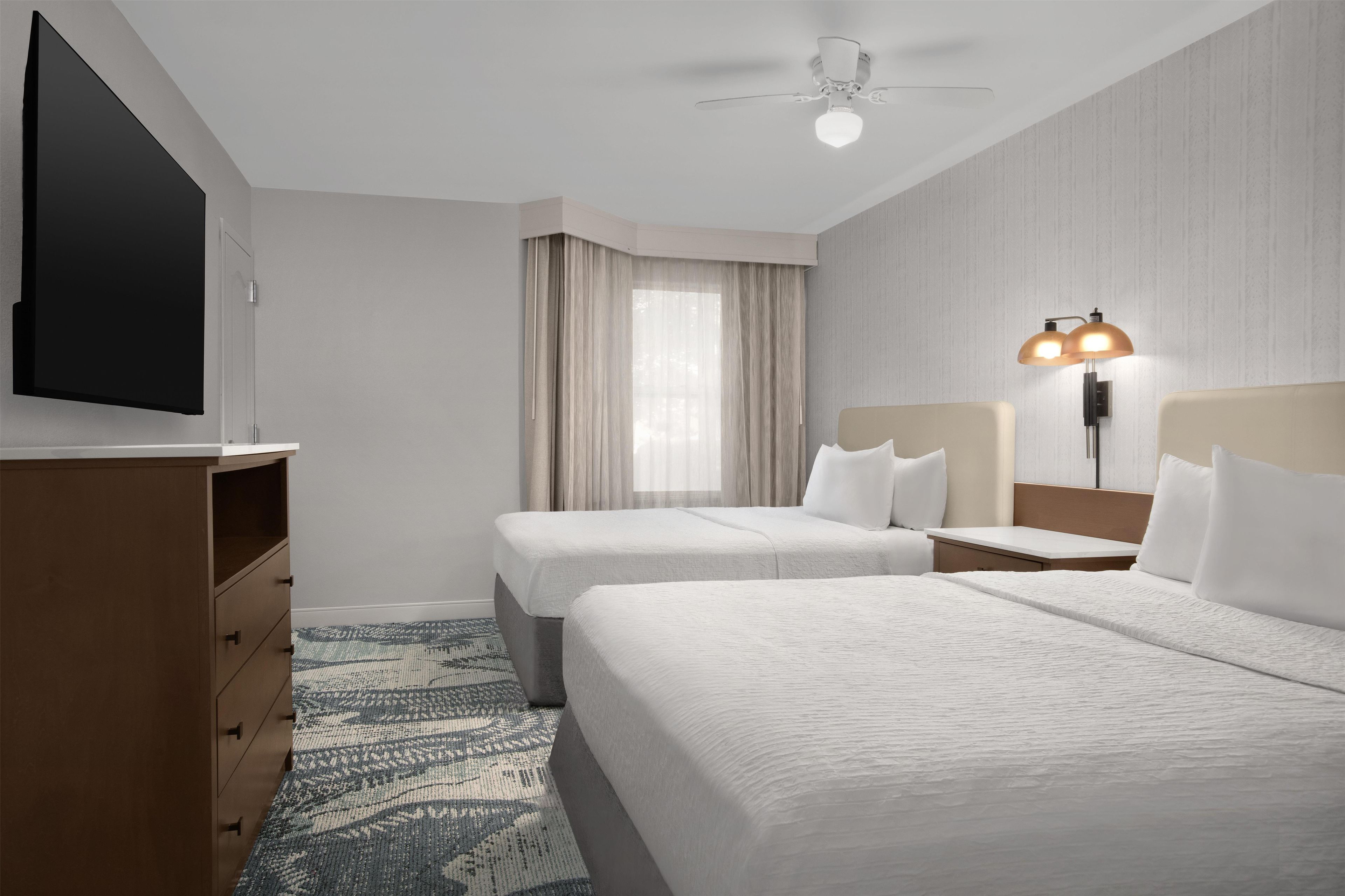 Habitación de hotel con dos camas, un televisor, ventilador de techo y mesita de noche con lámpara.