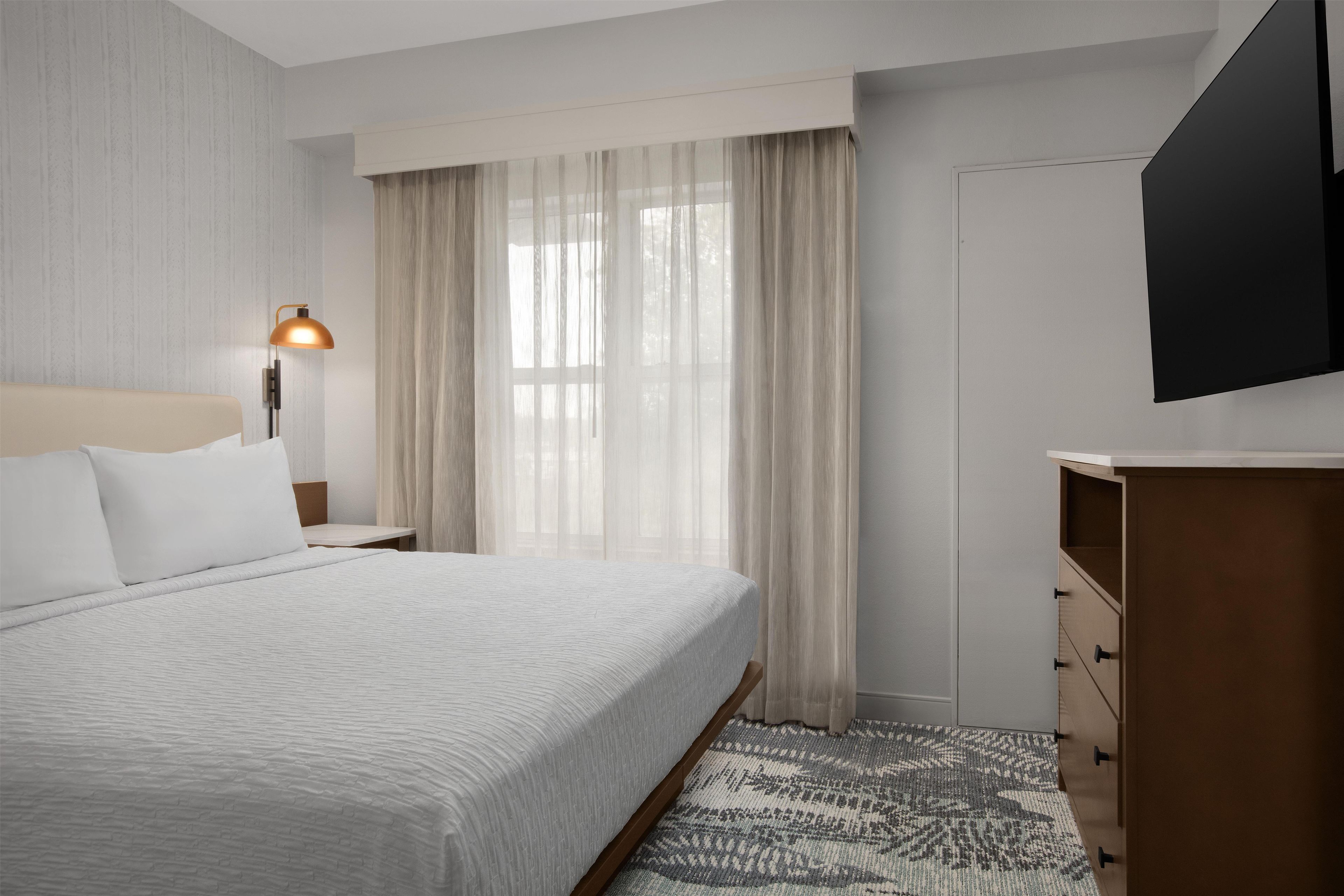 Habitación de hotel con una cama grande, una lámpara de noche, un televisor montado en la pared y una ventana con cortinas translúcidas.