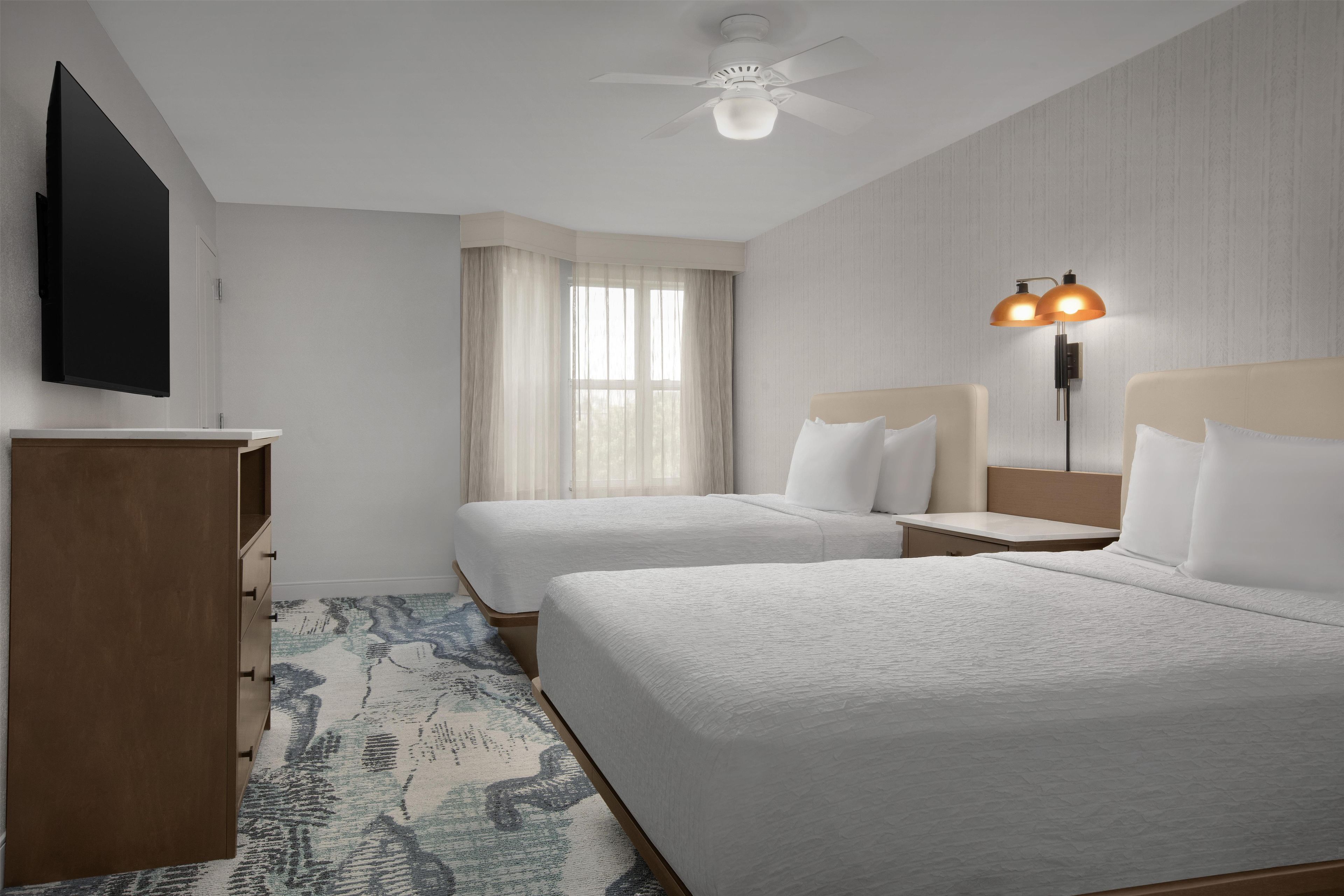 A hotel room with two beds, a flat-screen TV, a ceiling fan, a window with curtains, and a floor lamp.
