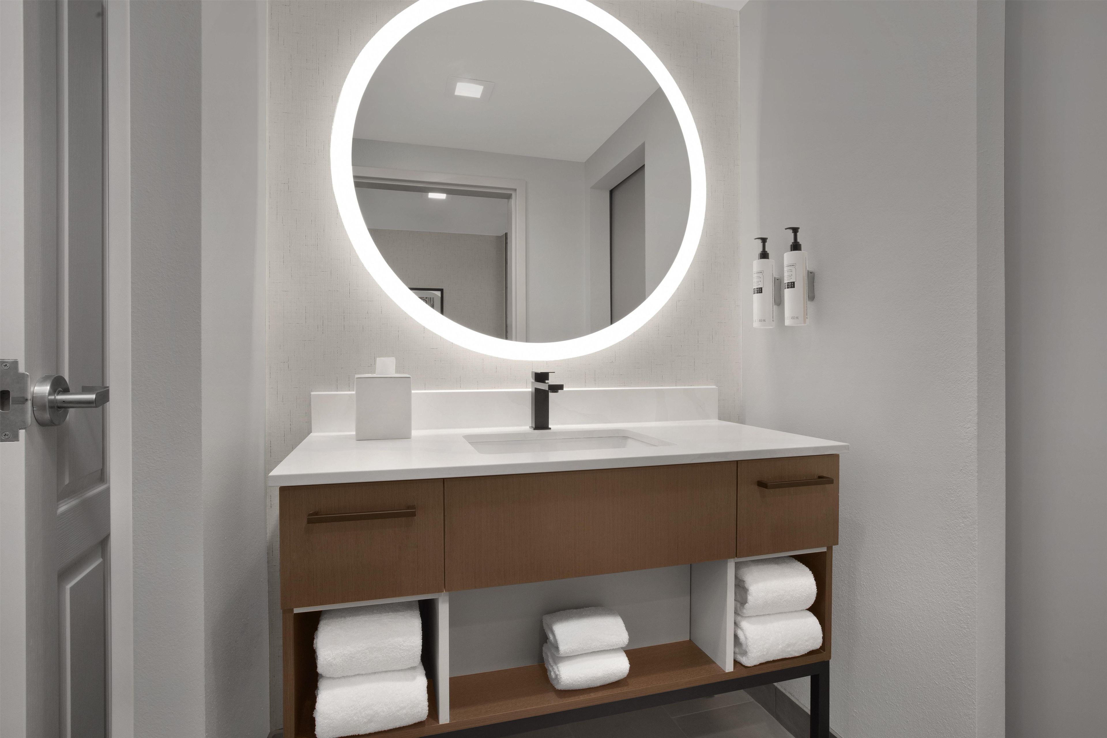 A modern bathroom with a round mirror, white sink, wooden vanity, and neatly folded towels.