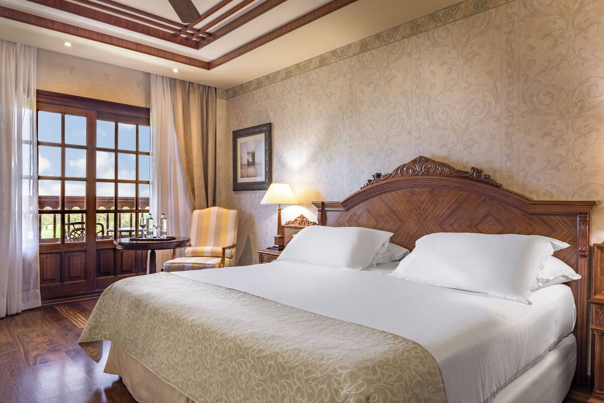 Ein Hotelzimmer mit einem großen Bett, einem Holzkopfende, einem kleinen Tisch mit Lampe, einem Sessel und einem Fenster mit Vorhängen.