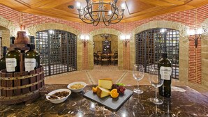 Une salle de dégustation de vins avec un mur en briques, un plafond en bois et un lustre. On y trouve des bouteilles de vin, du fromage, des raisins et des verres sur une table en marbre.