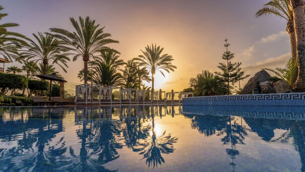 Une piscine dont l’eau reflète clairement les palmiers et où l’on aperçoit un coucher de soleil en arrière-plan.