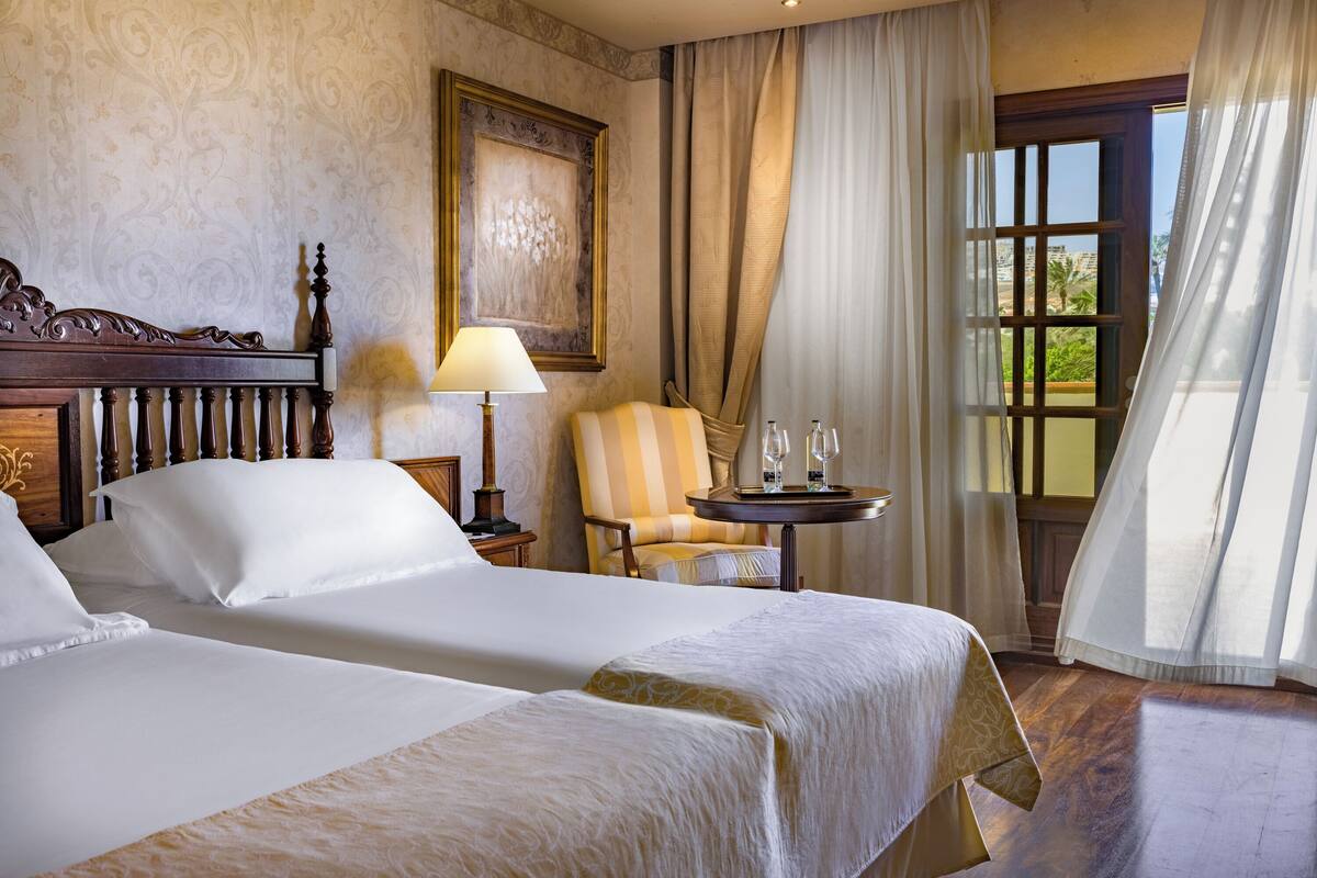 Ein Hotelzimmer mit zwei Betten, einem Sessel, einem Tisch mit Gläsern und einem Fenster mit Vorhängen.
