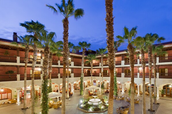 Un patio de hotel con palmeras, mobiliario exterior y una fuente central.