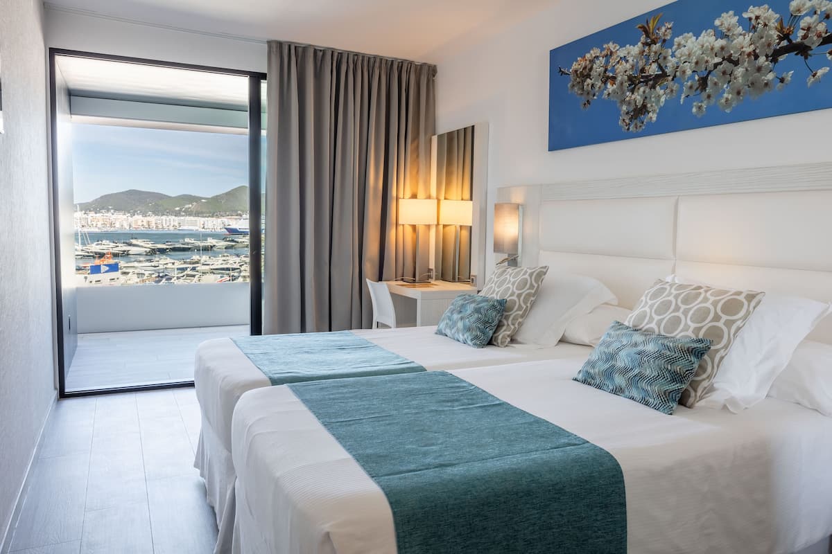 A hotel room with two beds, a desk, a chair, a lamp, and a view of a marina and cityscape.