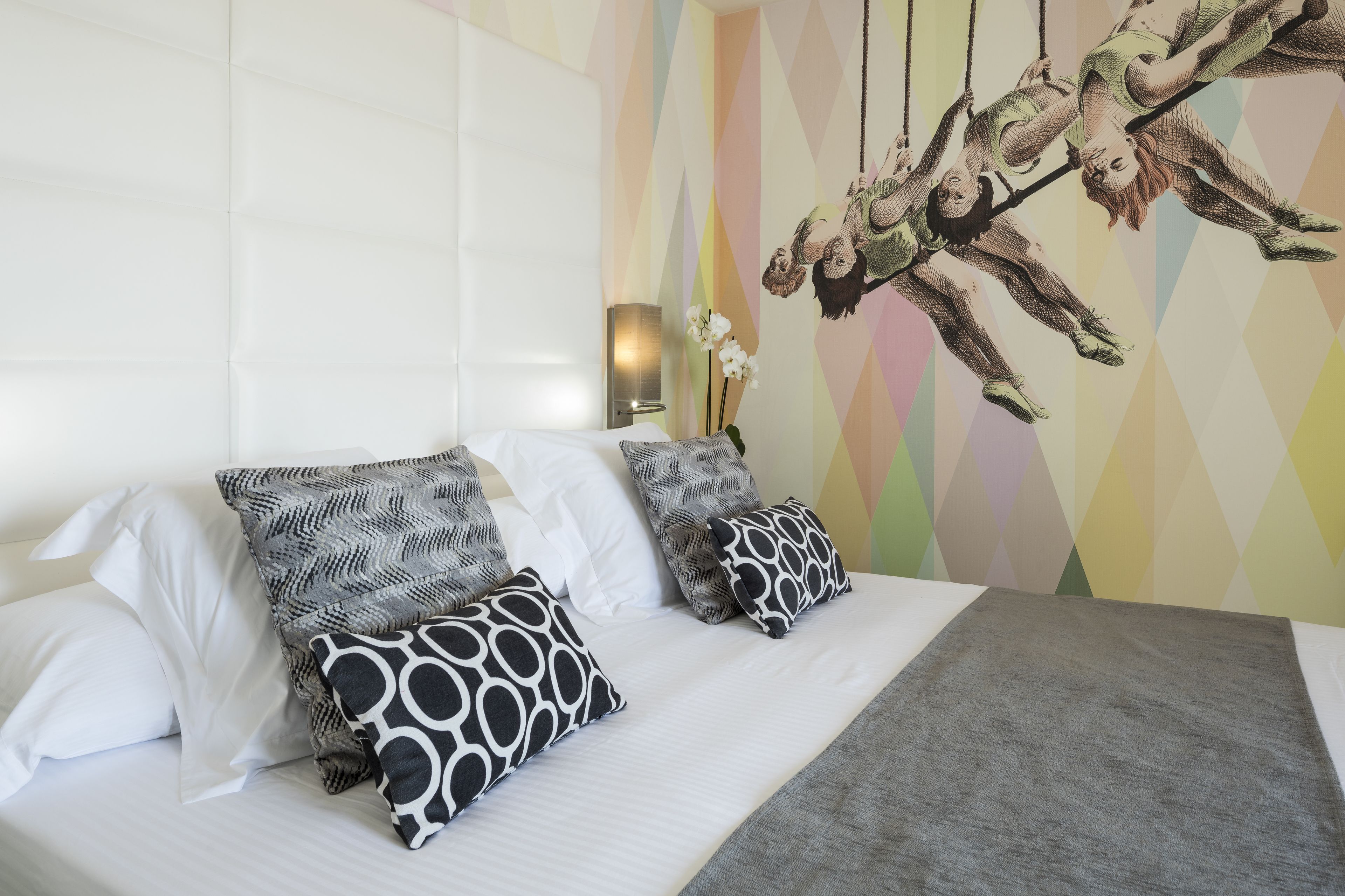 Una cama con almohadas blancas y estampadas, una colcha gris, y una pared con un mural geométrico y una escultura colgante.