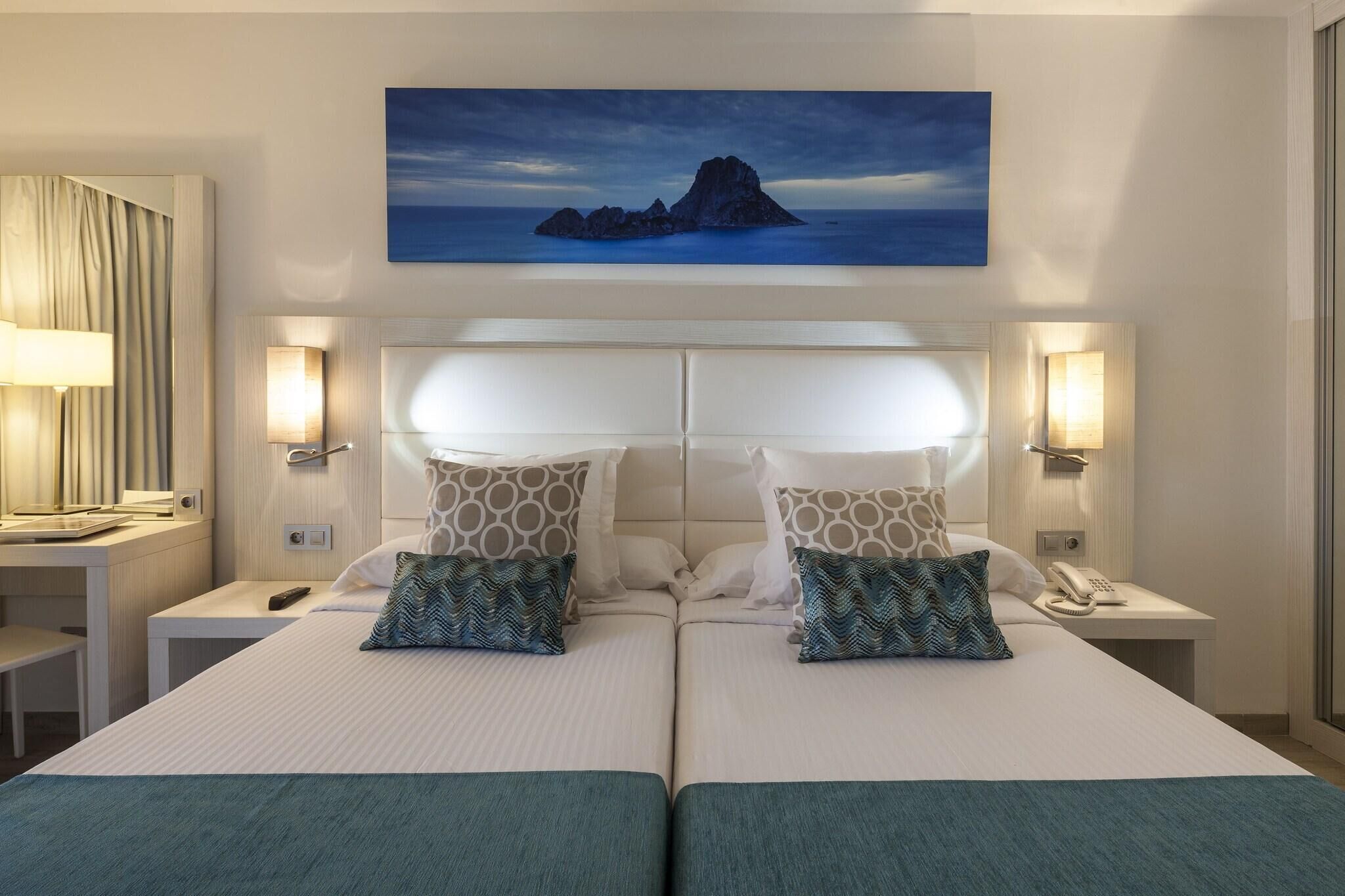A hotel room with a large bed, two bedside tables, a desk, a chair, a painting of a landscape, and a lamp.