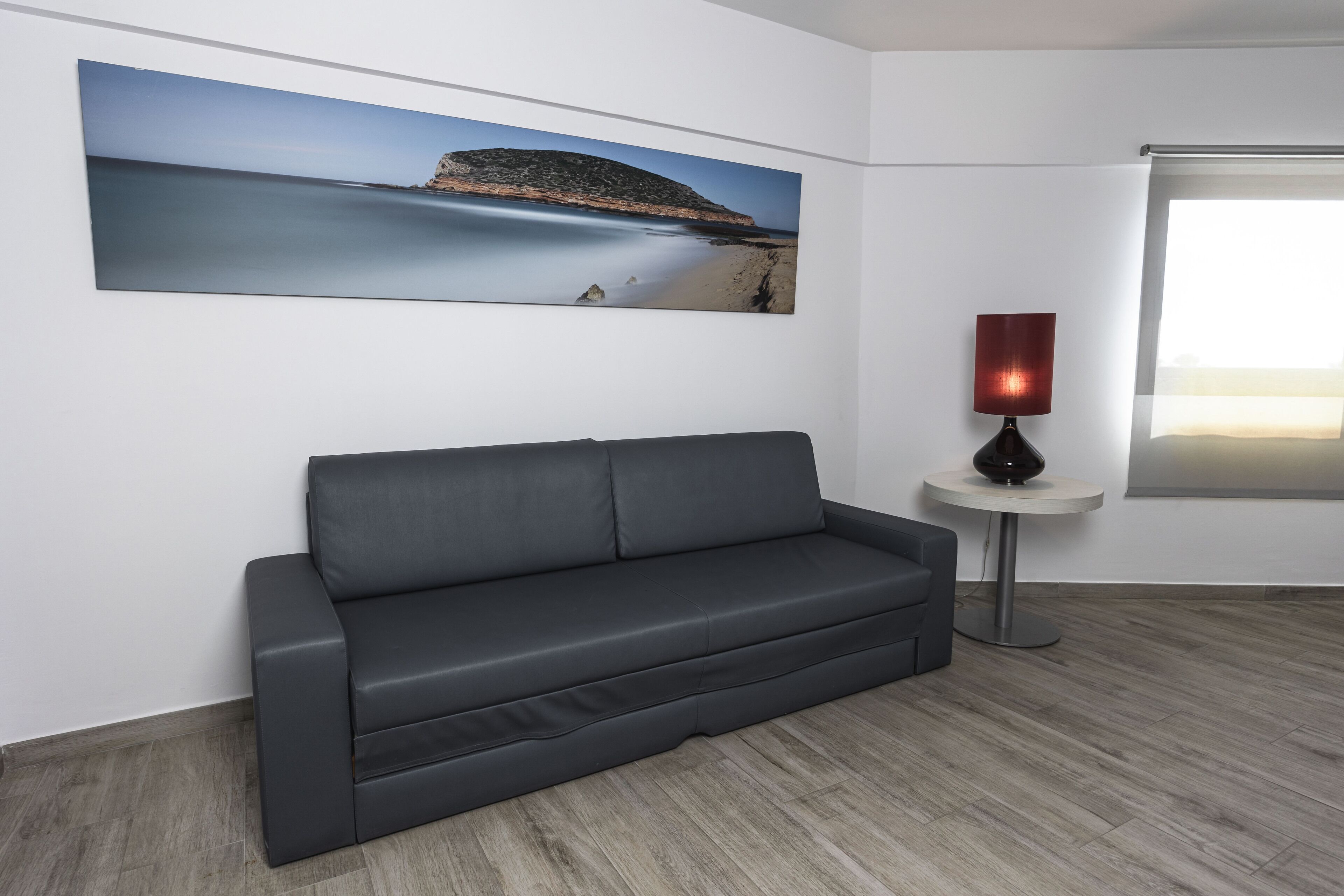 A modern living room with a grey sofa, a large landscape painting, a small round table with a lamp, and a large window.