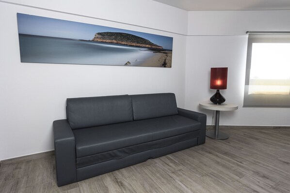 Una sala de estar moderna con un sofá gris, un cuadro de paisaje grande, una mesita redonda con una lámpara y un ventanal amplio.