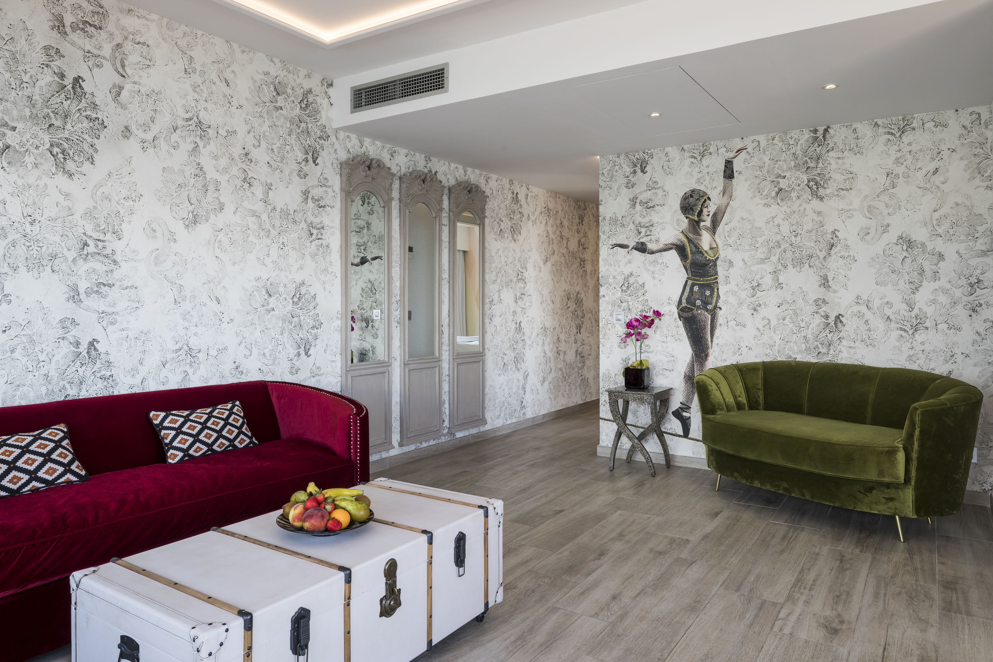 A modern living room with a red velvet sofa, a green velvet armchair, and a white coffee table with fruit.