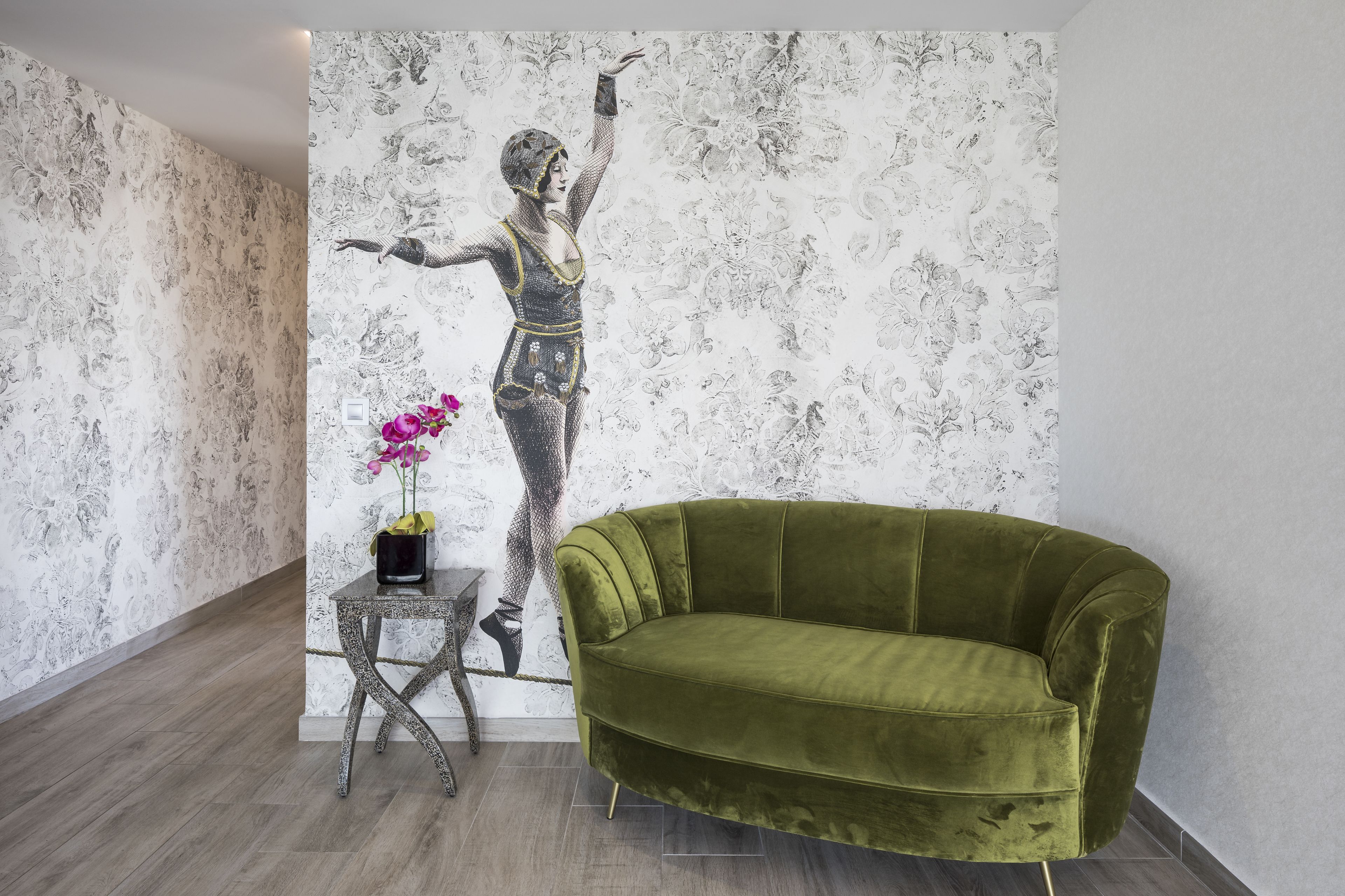 A room with a green velvet sofa, a small table with a vase of flowers, and a wall with a monochrome mural.
