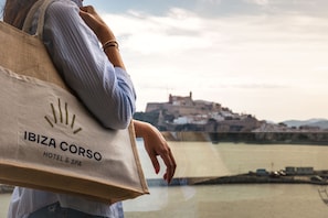 Una persona sosteniendo una bolsa con la leyenda "IBIZA CORSO HOTEL & SPA", observando un paisaje urbano costero.