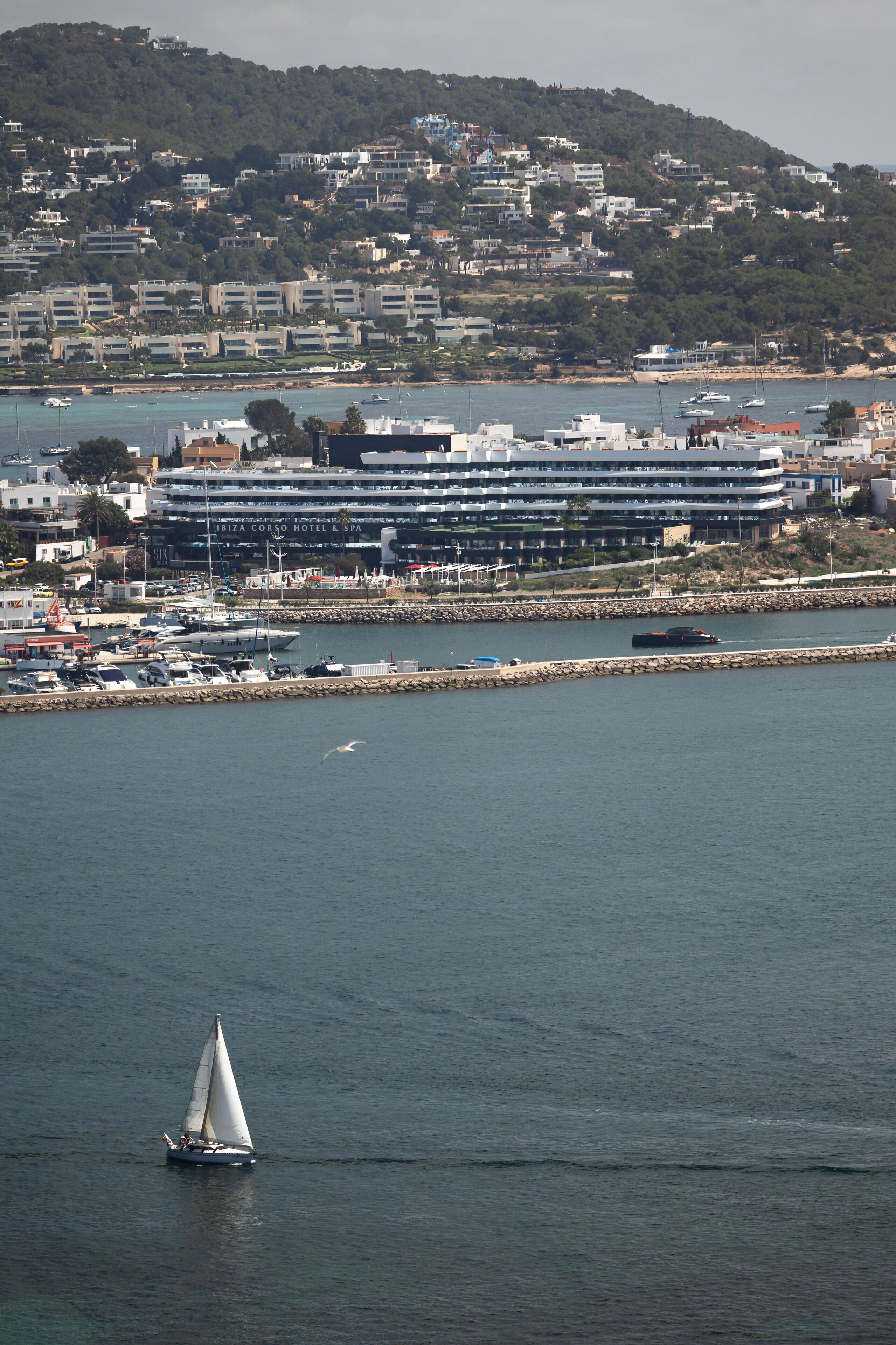 A sailboat on calm waters with a coastal town and a hotel in the background.