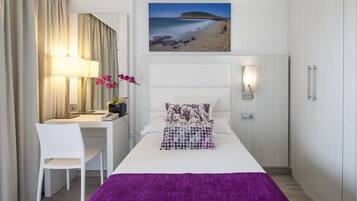 A hotel room with a bed, a desk, a chair, a lamp, a wall-mounted TV, and a painting of a beach.