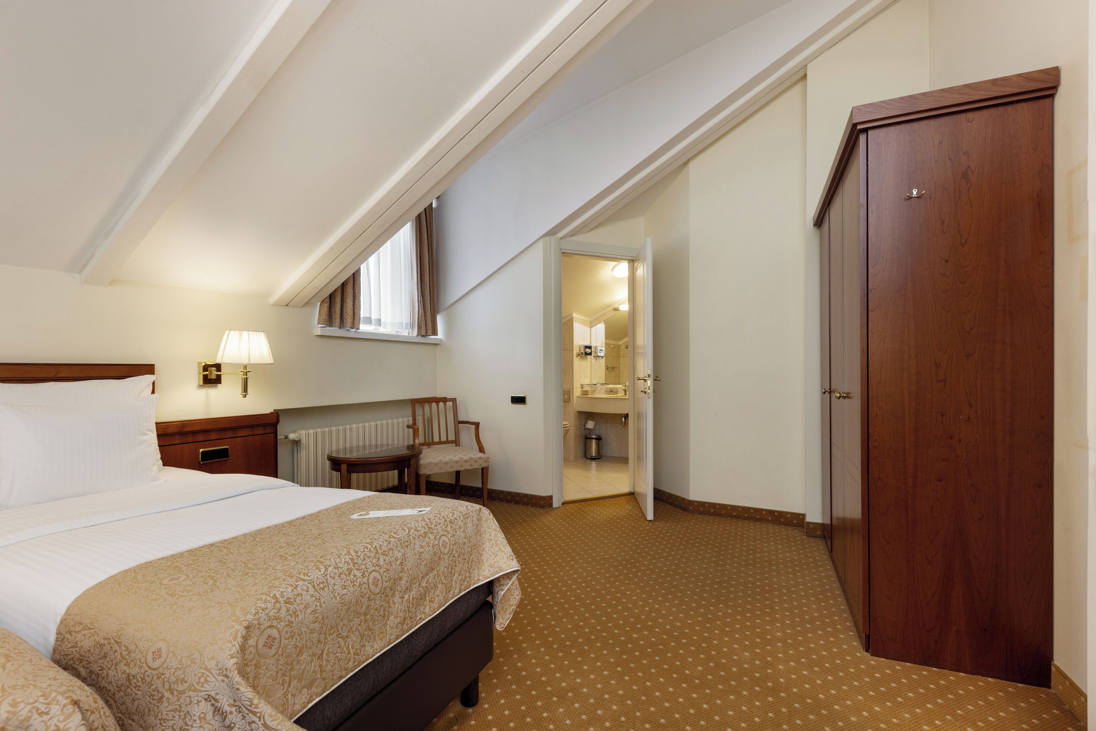 Une chambre d’hôtel avec un lit, un bureau, une chaise, une armoire et une salle de bain visible à travers une porte ouverte.