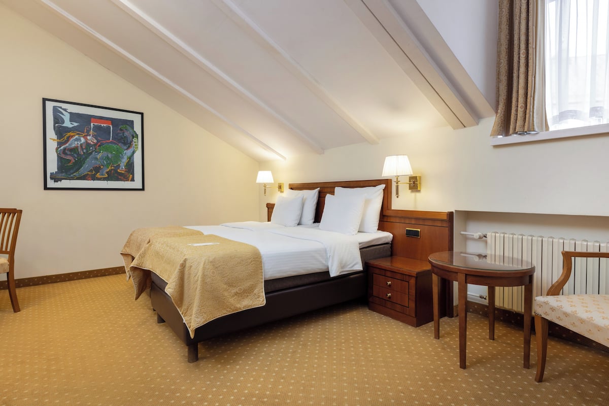 Ein Hotelzimmer mit Bett, Nachttischen, einem Sessel sowie einem Bild an der Wand und einem Fenster mit Vorhängen.