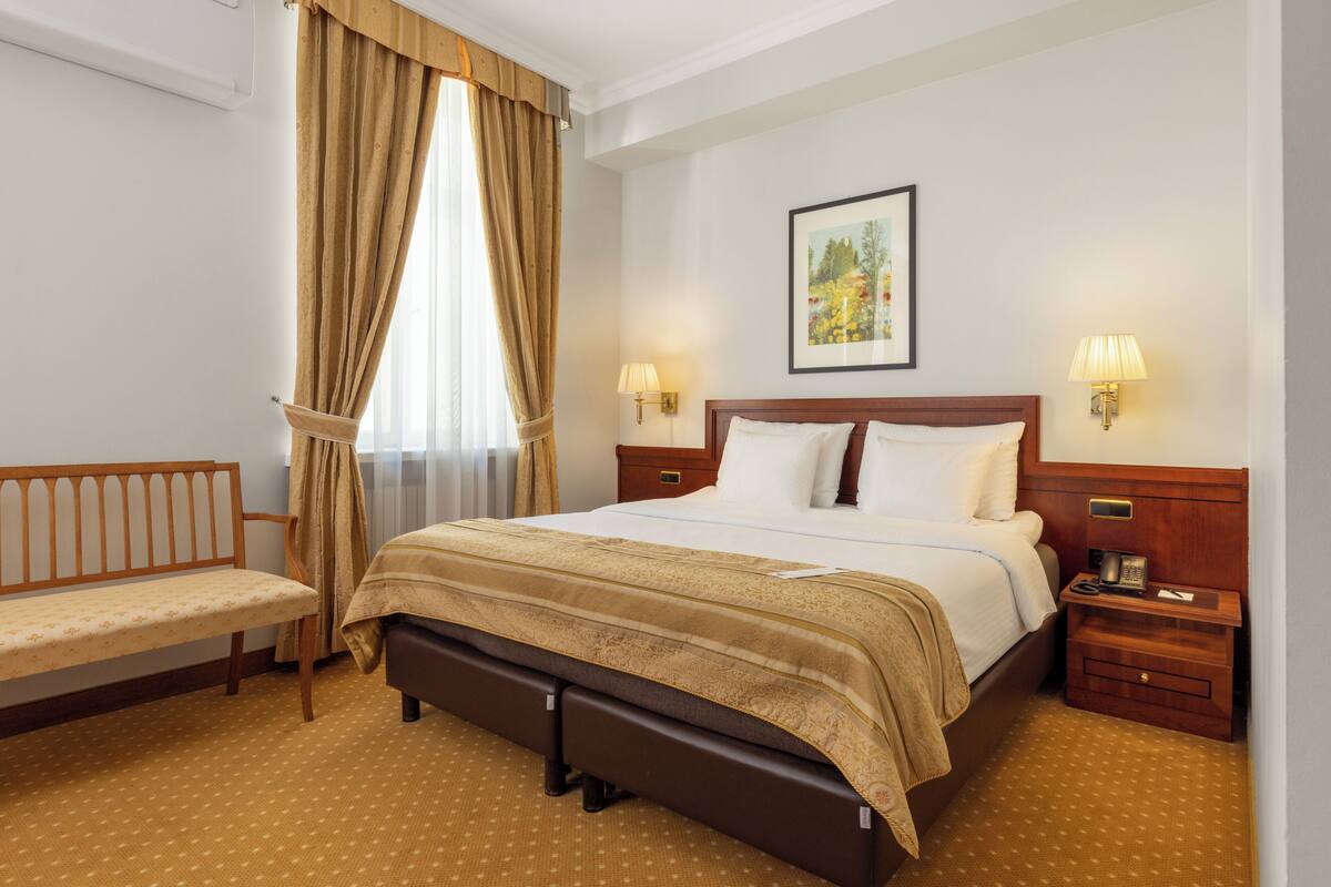Ein Hotelzimmer mit Bett, Nachttischen, einer Bank und einem Fenster mit Vorhängen.