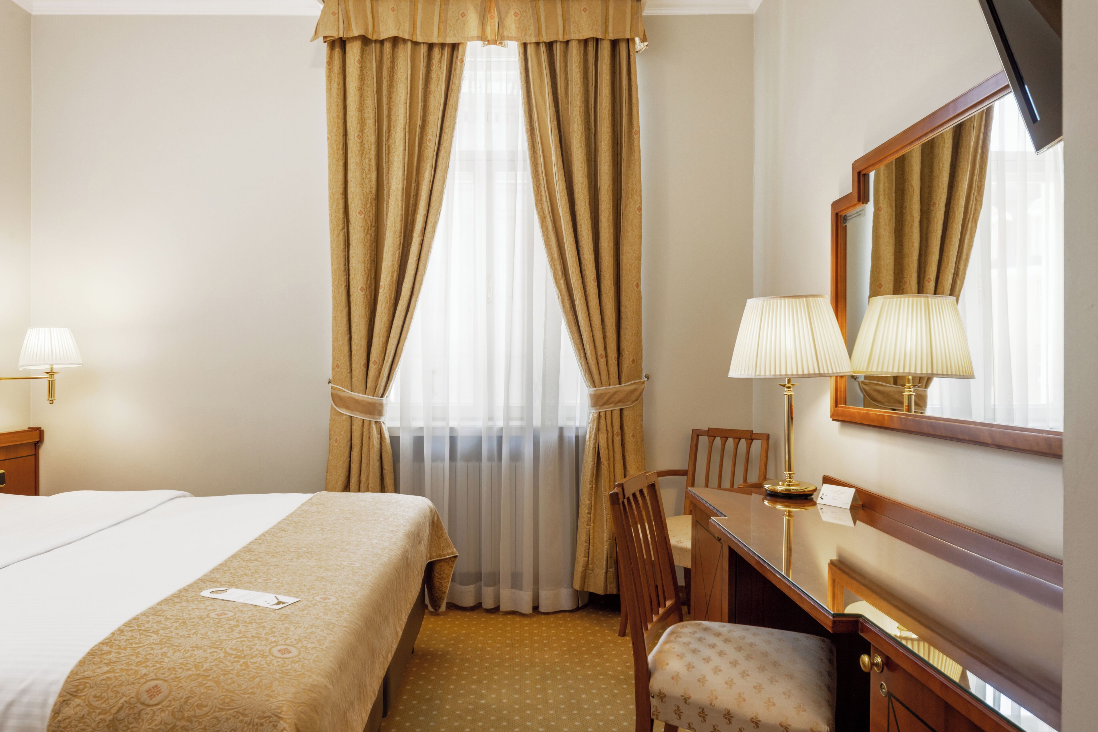 Une chambre d’hôtel avec un lit, un bureau et une chaise, un miroir et une fenêtre avec des rideaux.