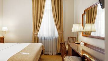 Une chambre d’hôtel avec un lit, un bureau et une chaise, un miroir et une fenêtre avec des rideaux.