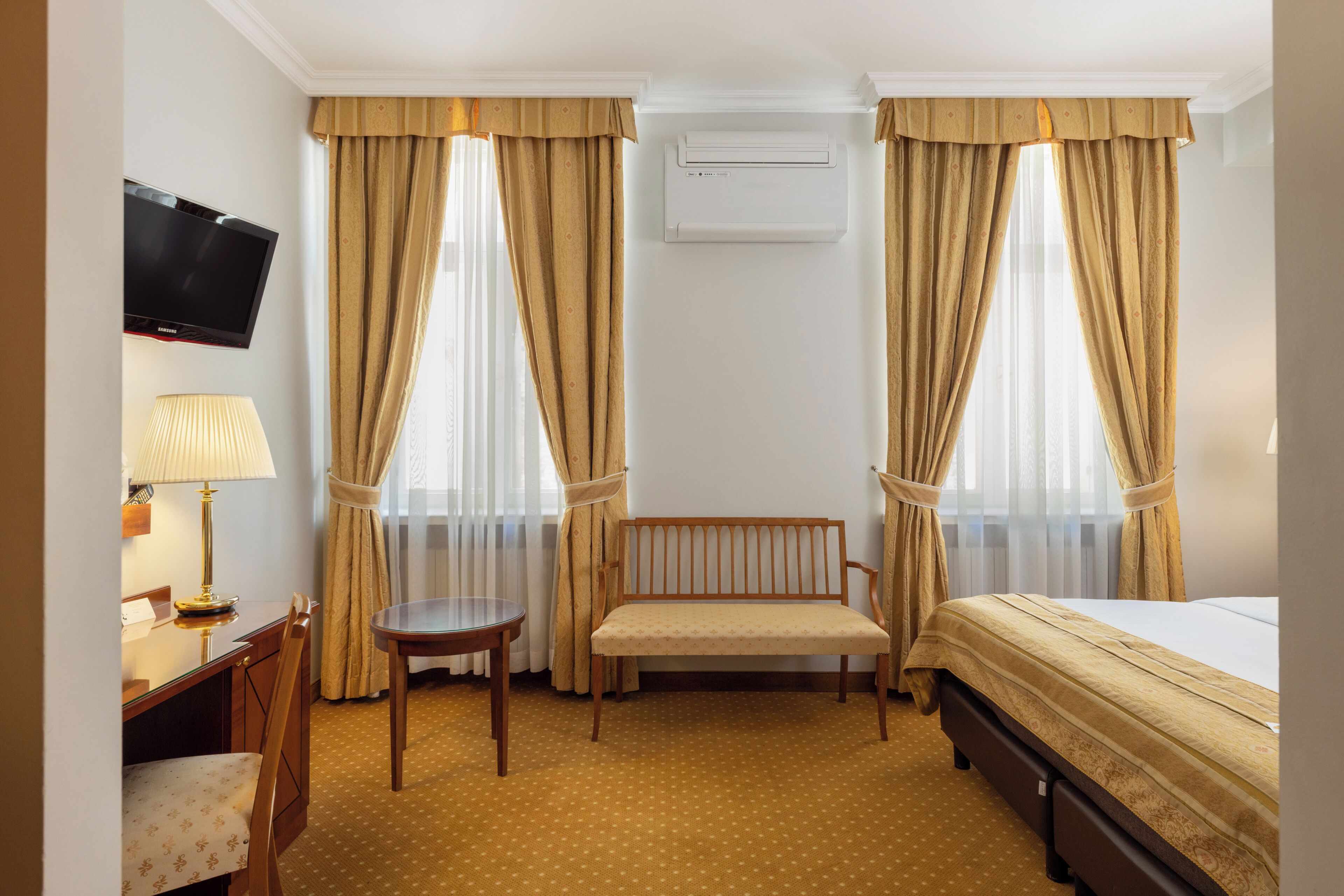 Habitación de hotel con cama, escritorio con lámpara, una silla, un banco y televisión en la pared.
