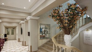 A hotel lobby with a tiled floor, a staircase with a white balustrade, a large white vase with flowers, and a display of bottles on shelves. - My City Hotel (Tallinn)